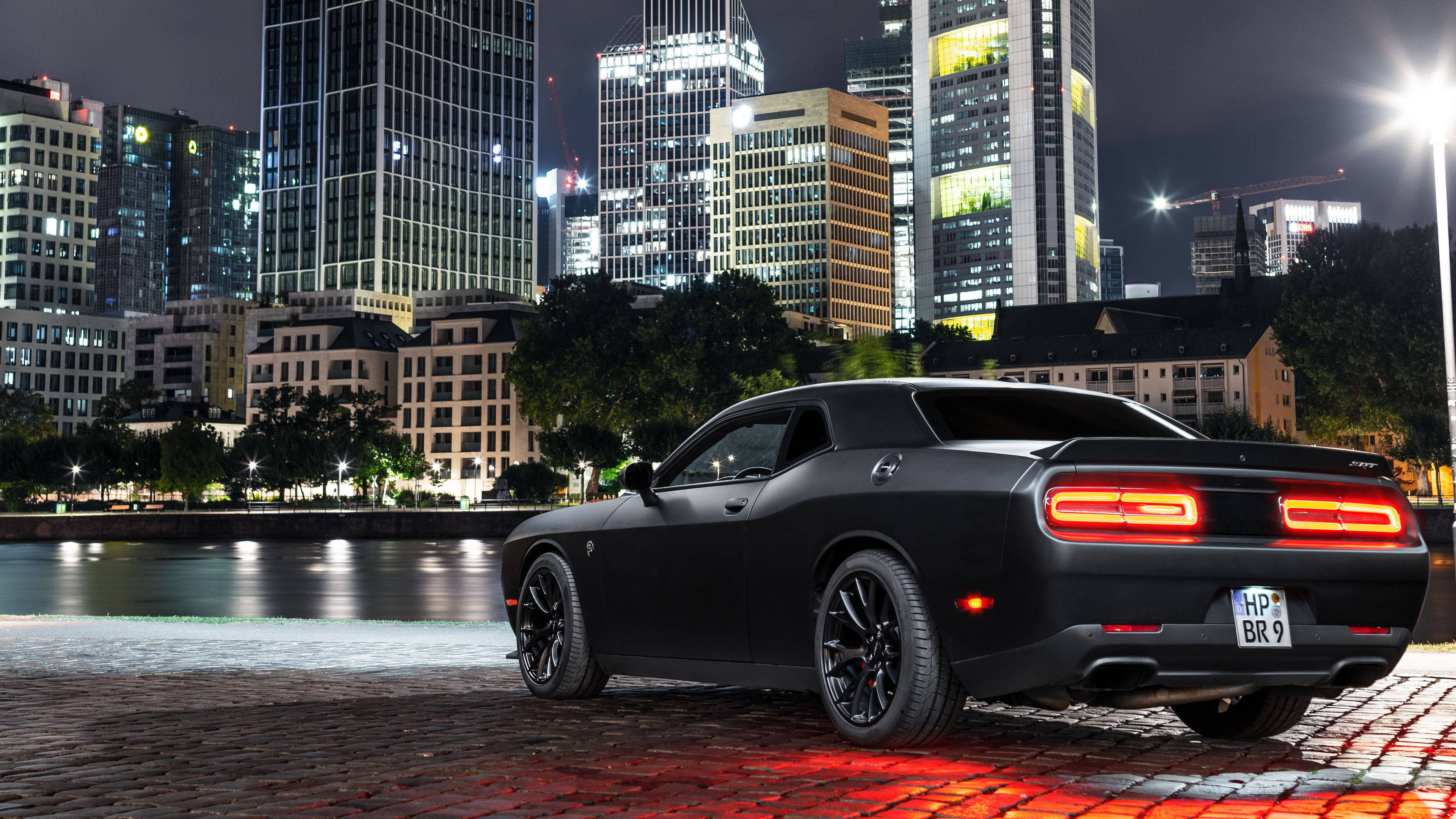 道奇挑戰者 桌布 壁紙 — 一輛消光黑色Dodge Challenger在夜晚停泊在鵝卵石海濱，背後是閃爍的城市天際線。