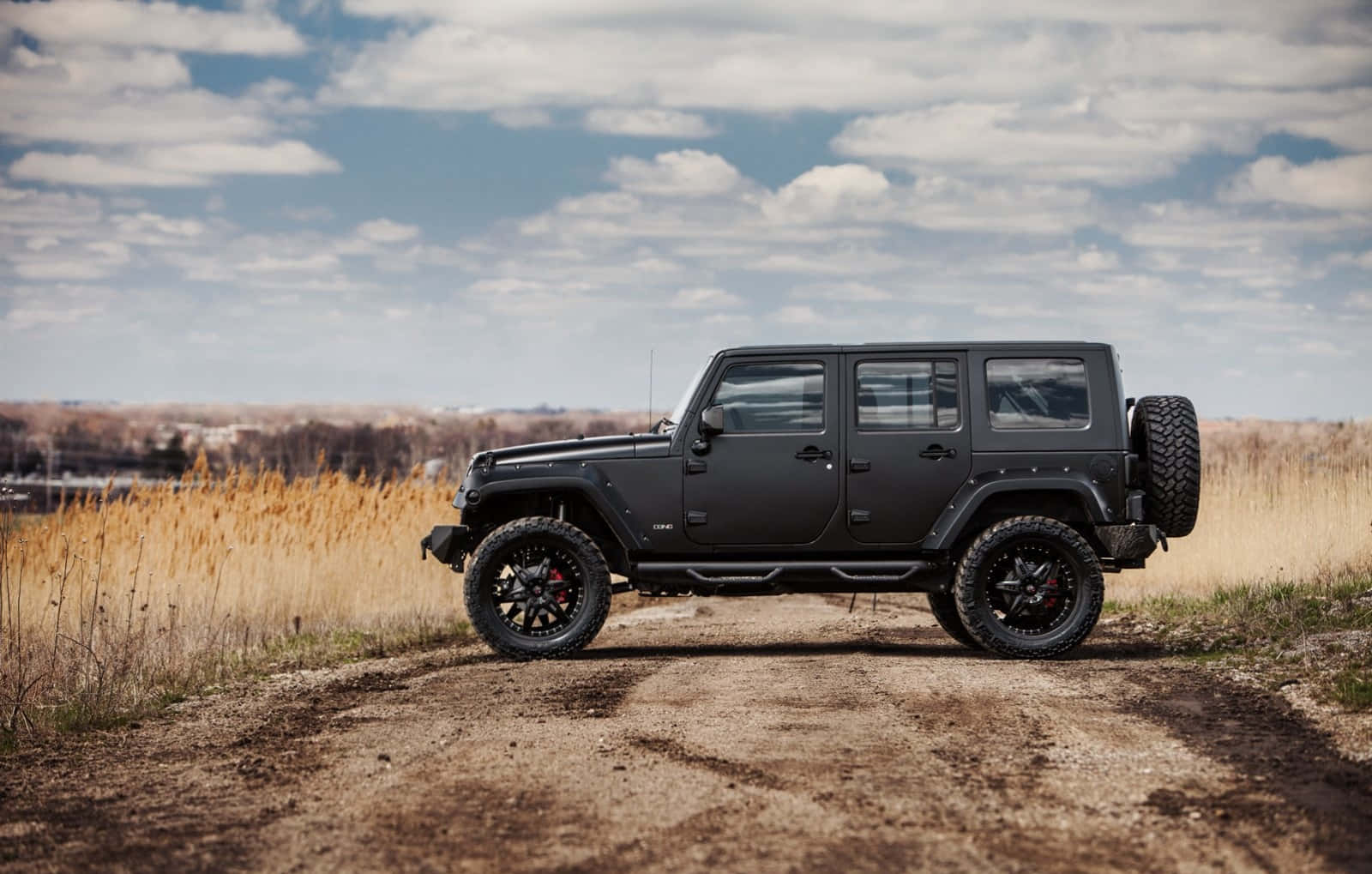 黑色吉普車 桌布 壁紙 — 一輛消光黑色Jeep Wrangler停在穿過金色草叢的泥土小徑上，天空部分多雲。