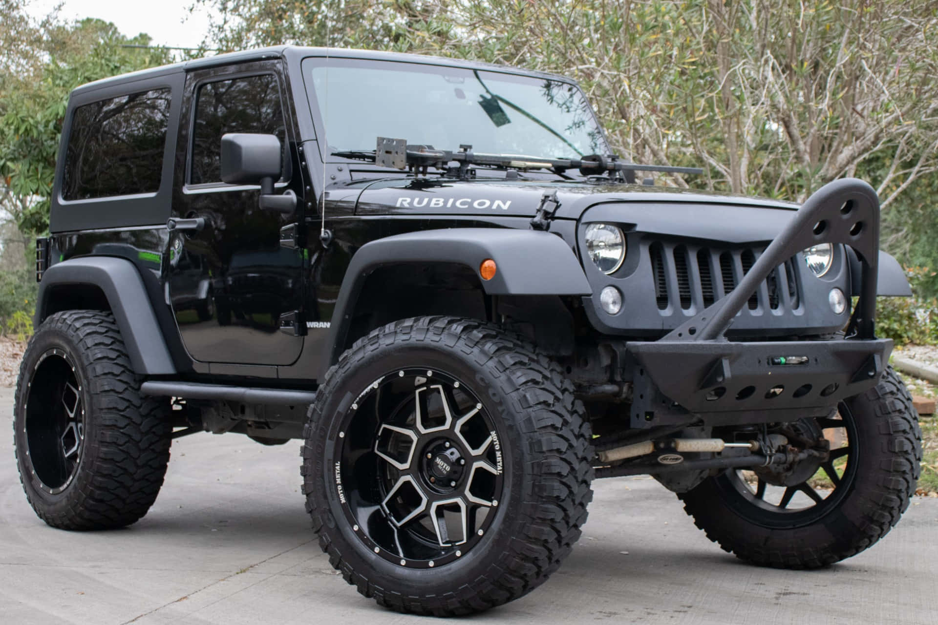 Jeep Wrangler 桌布 壁紙 — 一輛消光黑色Jeep Wrangler Rubicon配備超大輪胎和客製化輪圈，停放在乾樹環繞的混凝土表面上。