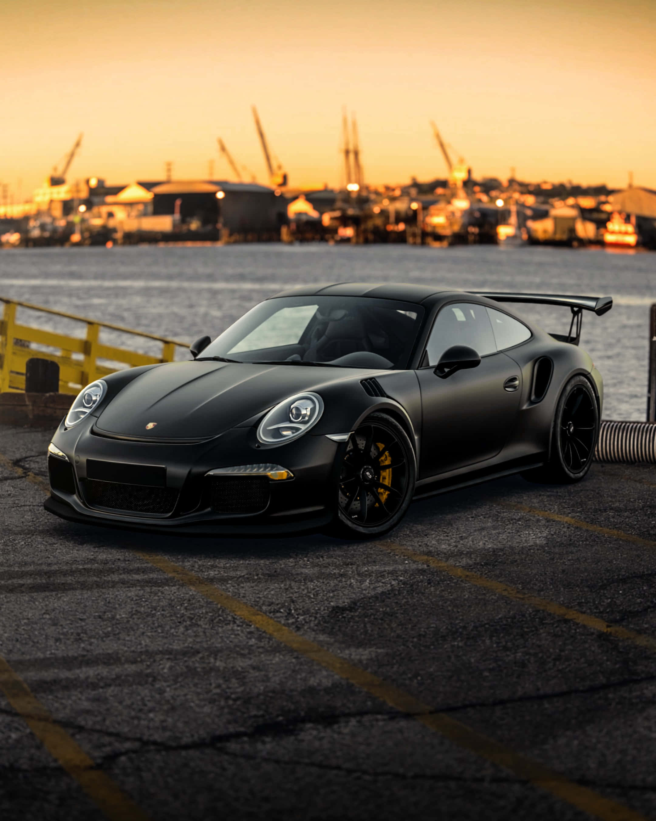 911 iPhone 桌布 壁紙 — 一輛消光黑色保時捷911 GT2 RS在黃金時刻停泊在碼頭，背景有起重機和水。