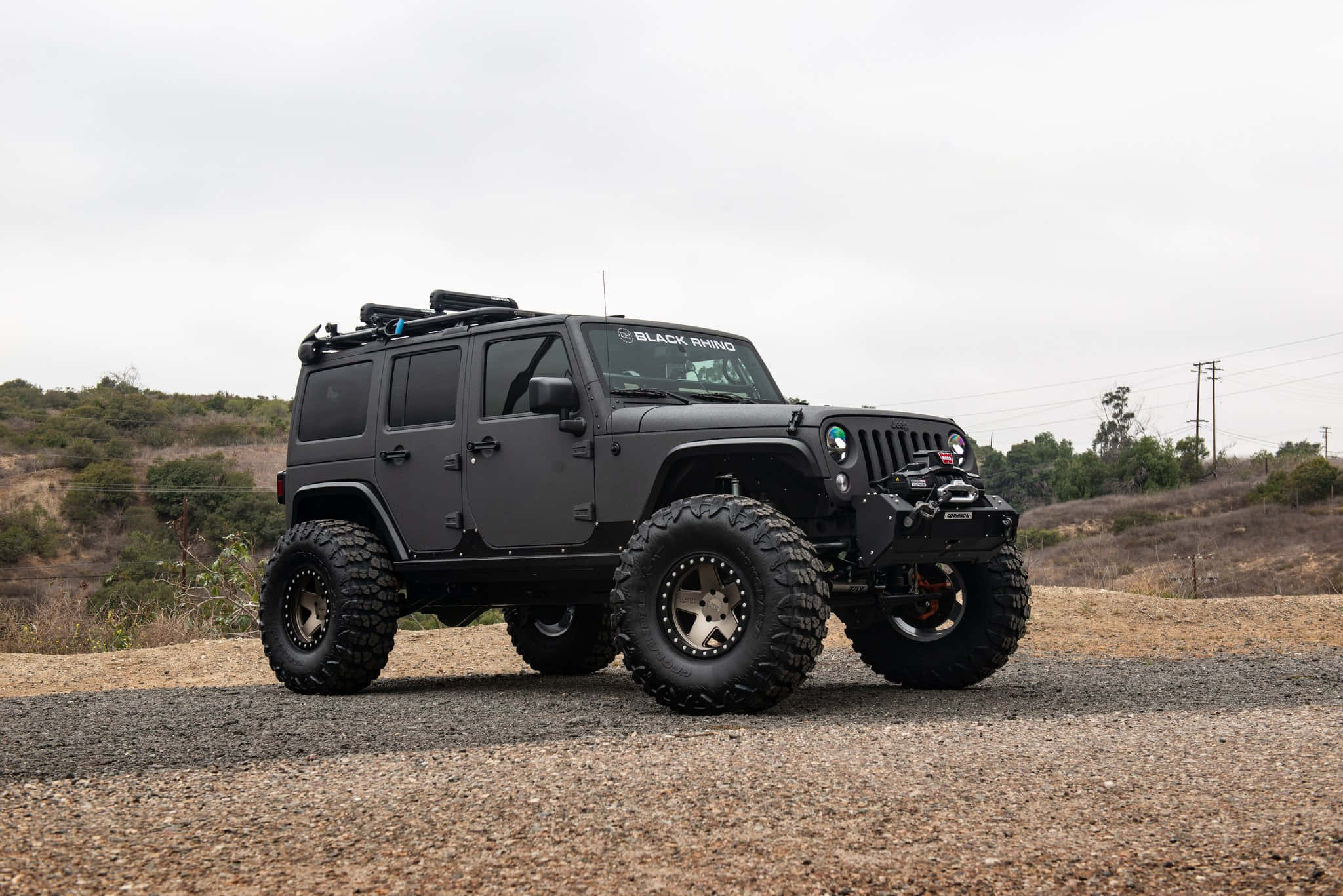 黑色吉普車 桌布 壁紙 — 一輛消光黑色升高Jeep Wrangler配備超大輪胎，停在碎石路上，背景是朦朧多雲的天空。