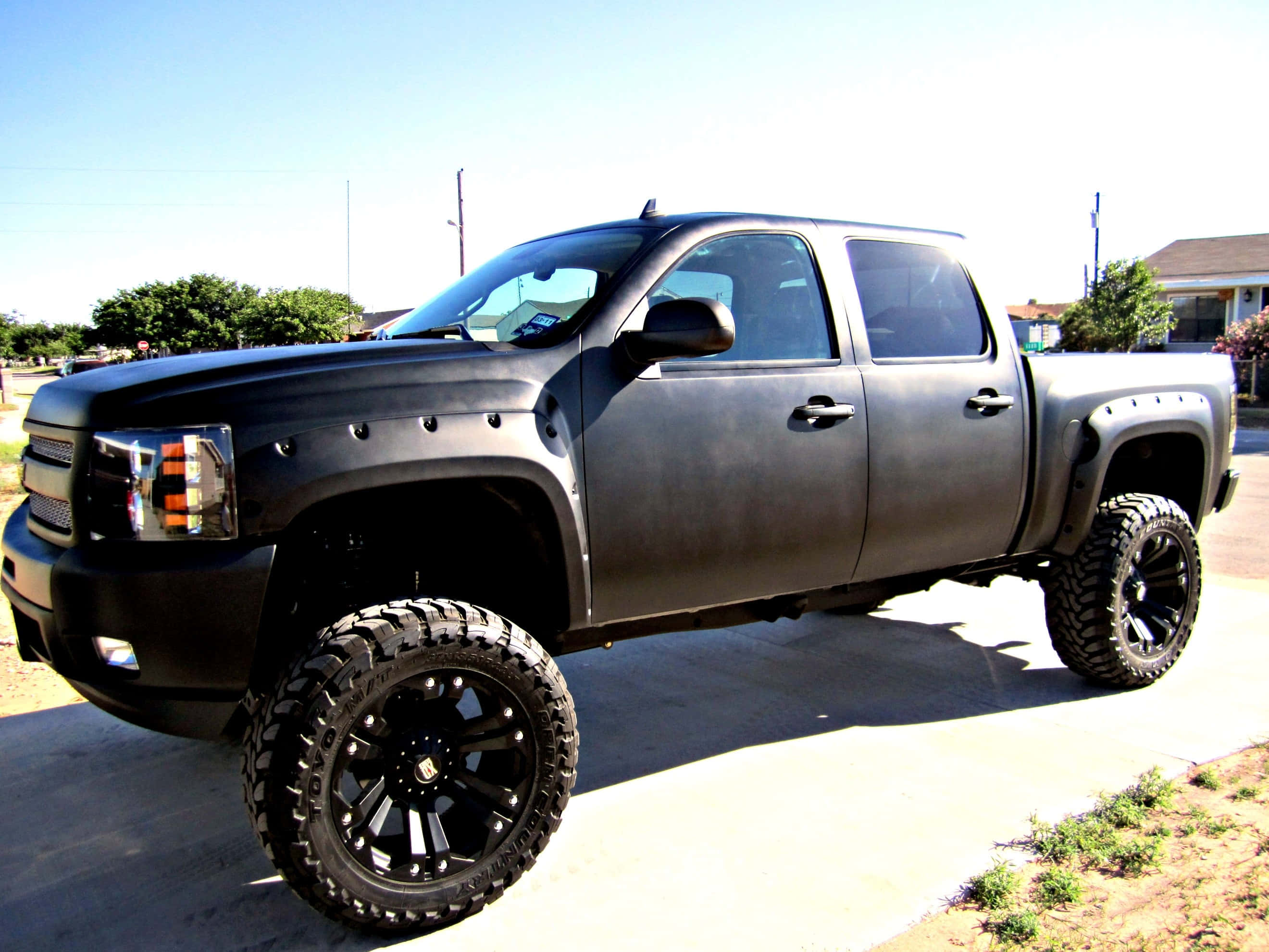 Jacked Up Trucks 桌布 壁紙 — 一輛消光黑色升高底盤的雪佛蘭 Silverado 皮卡車，配備超大輪胎，在明亮的日光下停在混凝土車道上。