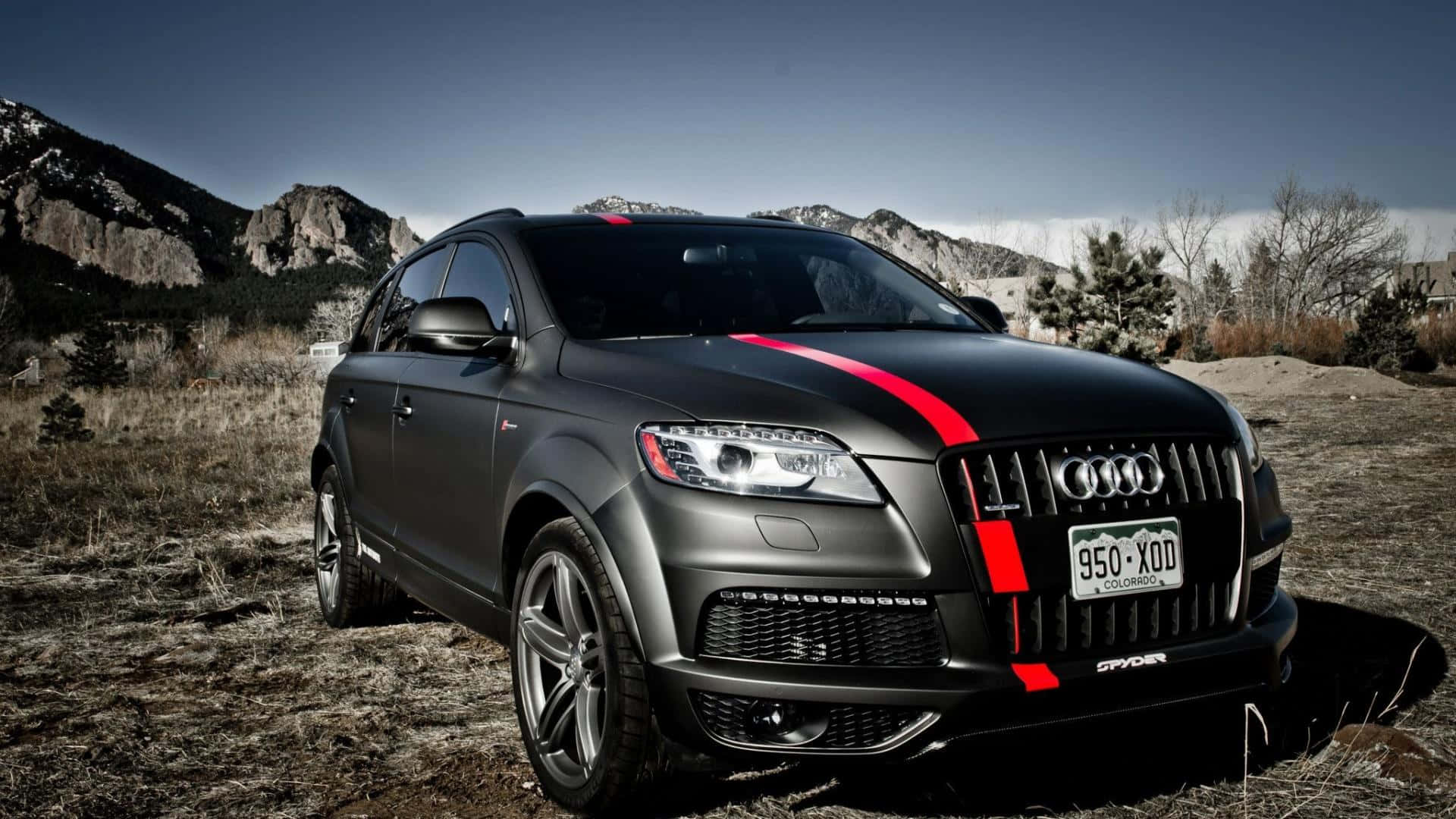 Audi Q7 桌布 壁紙 — 一輛消光黑色奧迪Q7，帶有醒目的紅色賽車條紋，停在乾燥的草地上，背景是岩石山脈和清澈的藍天。