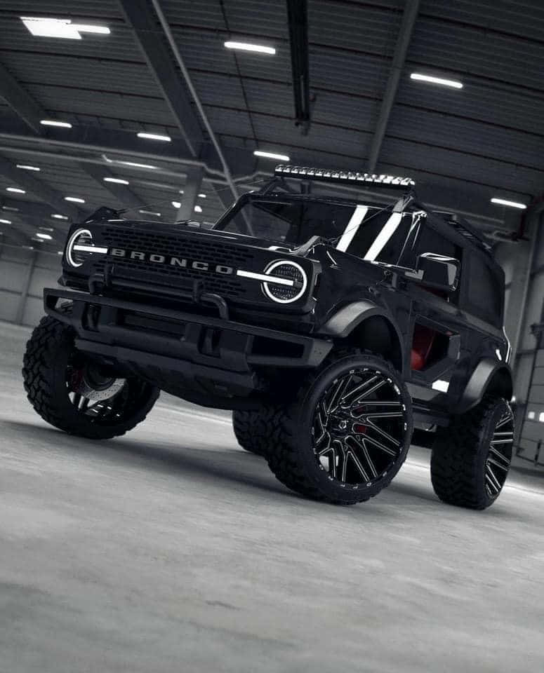 Off-road Car 桌布 壁紙 — 一輛消光黑色客製化Bronco SUV，配備超大輪圈和車頂燈，停放在寬敞的工業車庫中，上方有明亮的頂燈。