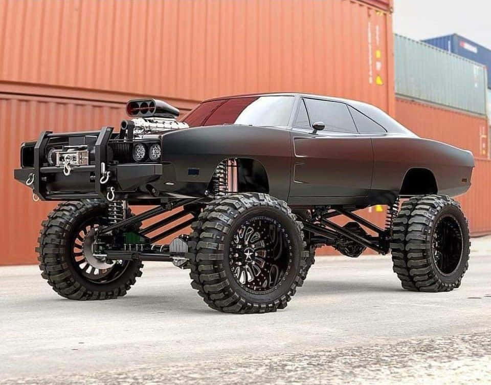 Off-road Car 桌布 壁紙 — 一輛消光黑色怪獸卡車，配備超大輪胎和裸露懸吊系統，停在堆疊的貨運貨櫃前。