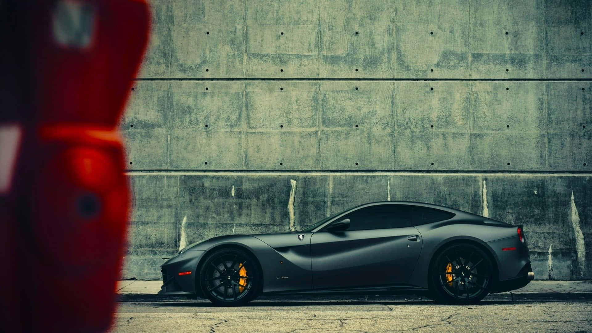 Ferrari F12 Berlinetta 桌布 壁紙 — 一輛消光黑色法拉利跑車停靠在風化的混凝土牆前，前景有一個模糊的紅色物體。