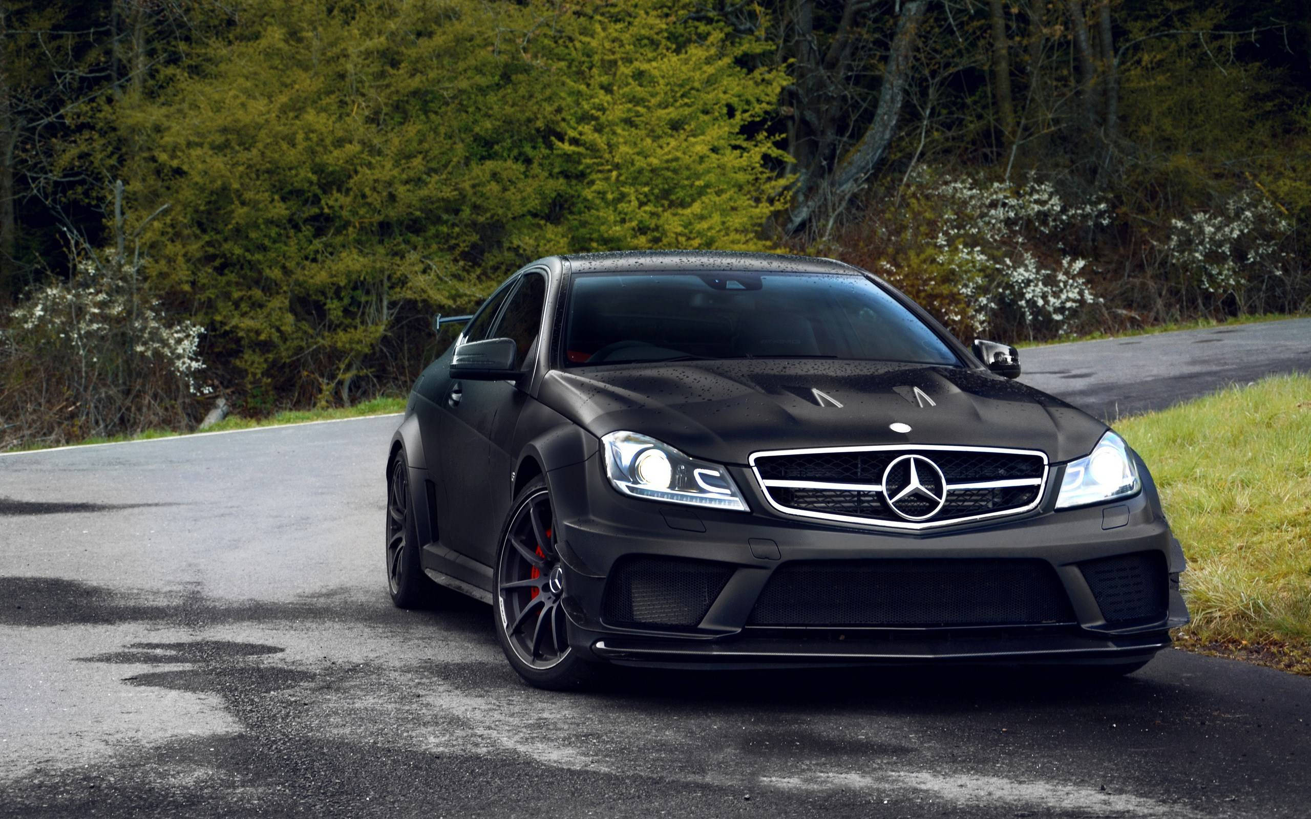 汽車 桌布 壁紙 — 一輛消光黑色的 Mercedes-AMG C63 停在潮濕的森林道路上，背景有綠色樹葉和白色花朵。