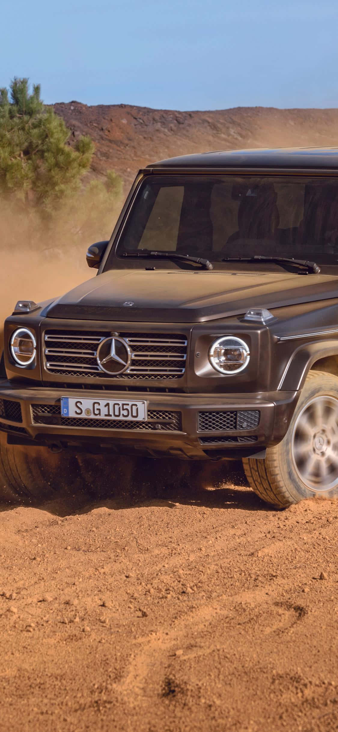桌布 壁紙 — 一輛消光黑色的 Mercedes G-Class SUV 在陽光明媚的沙漠景觀中行駛，揚起紅色塵土，周圍有岩石丘陵和稀疏的綠樹。