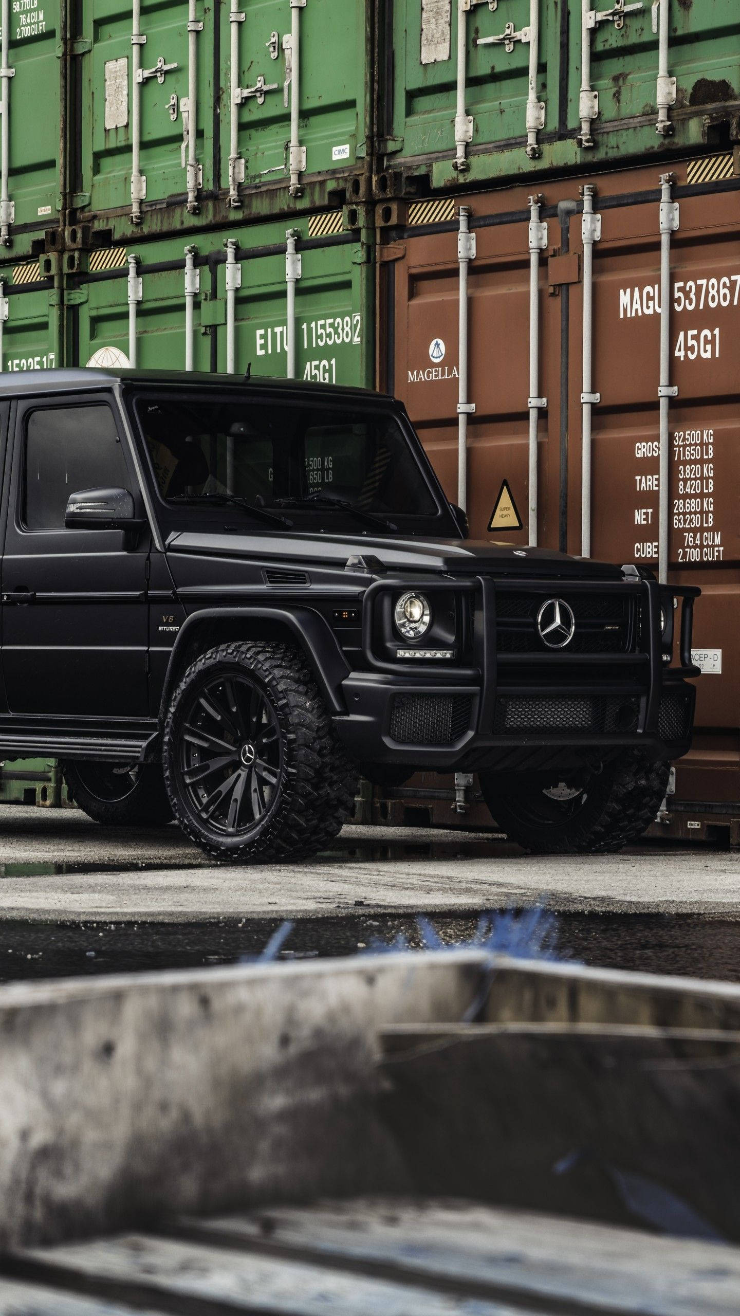 賓士 iPhone 桌布 壁紙 — 一輛消光黑色的 Mercedes G-Wagen 堅固地矗立在工業場地堆疊的綠色和棕色貨運貨櫃前。