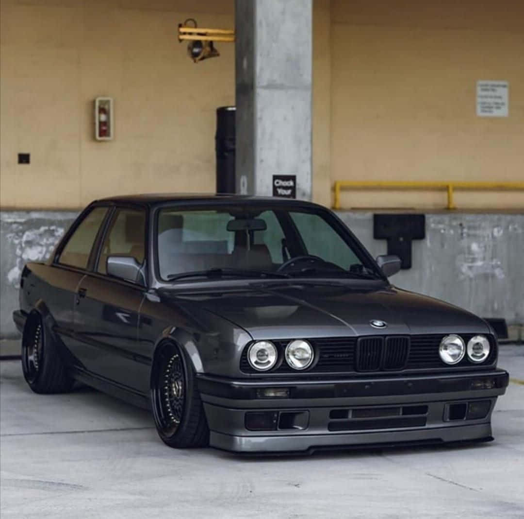 BMW 動圖 桌布 壁紙 — 一輛消光黑色的BMW E30雙門轎跑車配備客製化輪圈，在混凝土停車場的人造燈光下低趴停放。