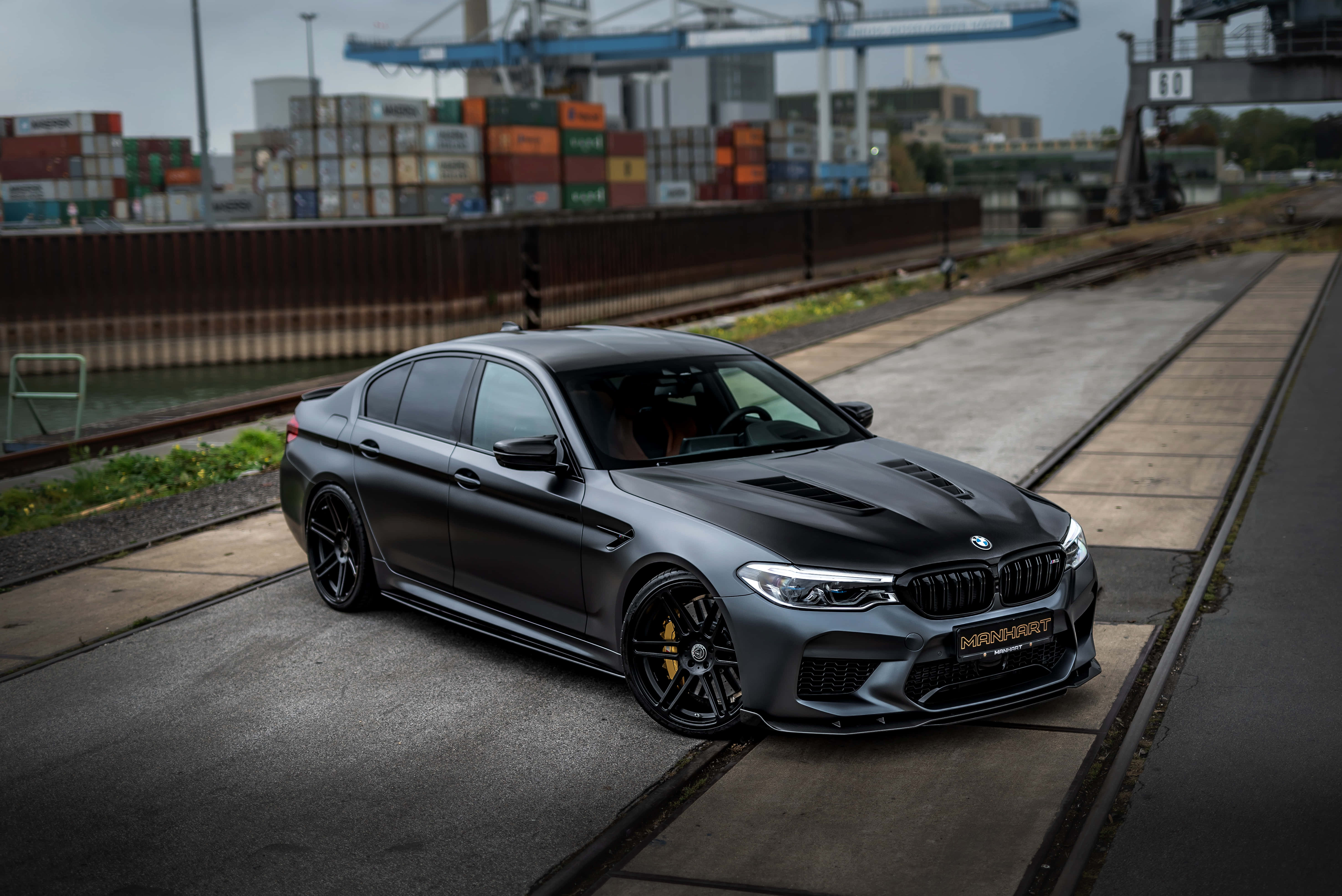 桌布 壁紙 — 一輛消光黑色的BMW M5跑車停放在鐵路軌道旁，背景有貨運貨櫃和起重機的工業場景。