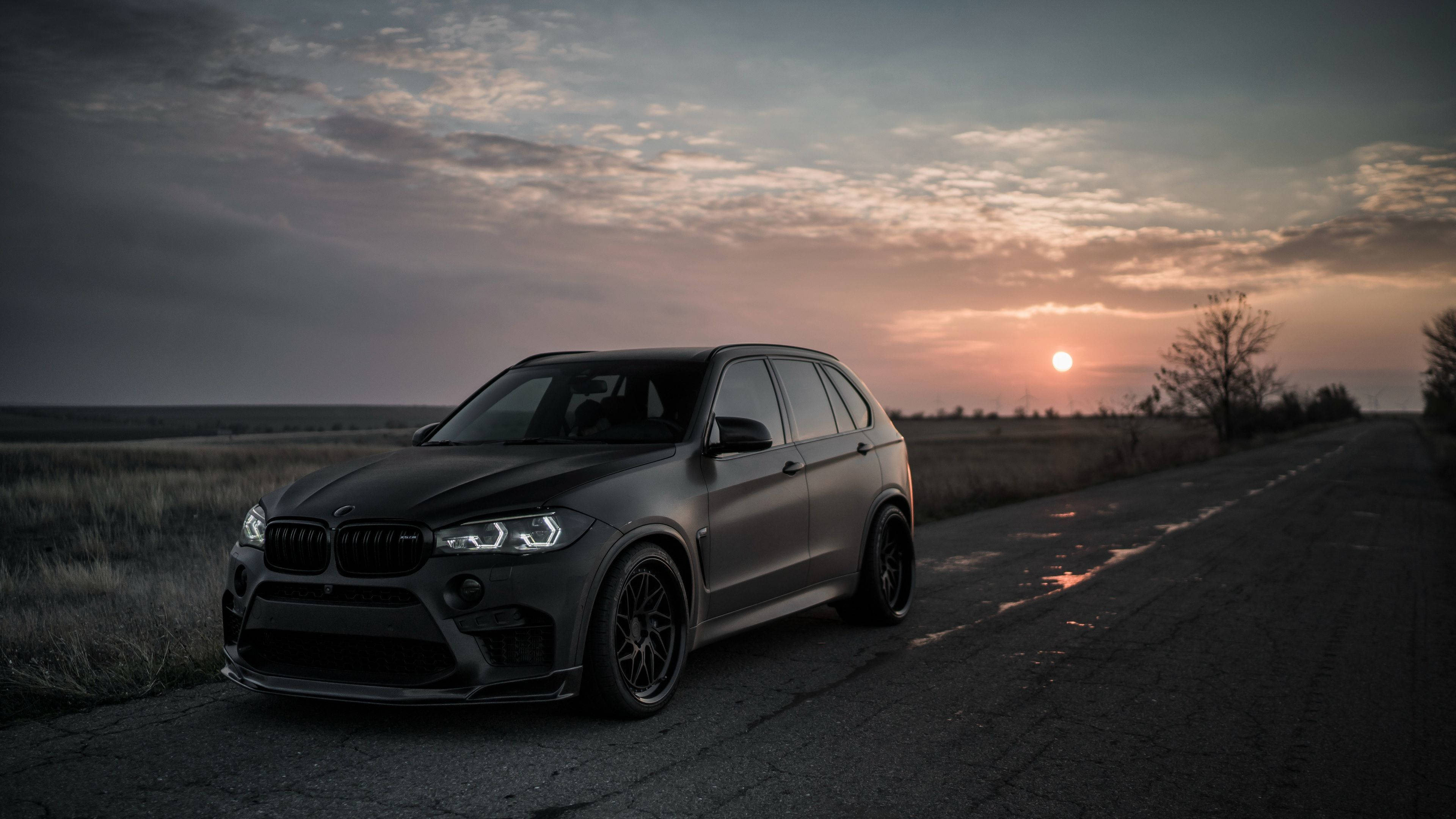 BMW 動圖 桌布 壁紙 — 一輛消光黑色的BMW X5 M停在潮濕的鄉村道路上，夕陽西下，天空有戲劇性的雲彩和溫暖的金色光芒。
