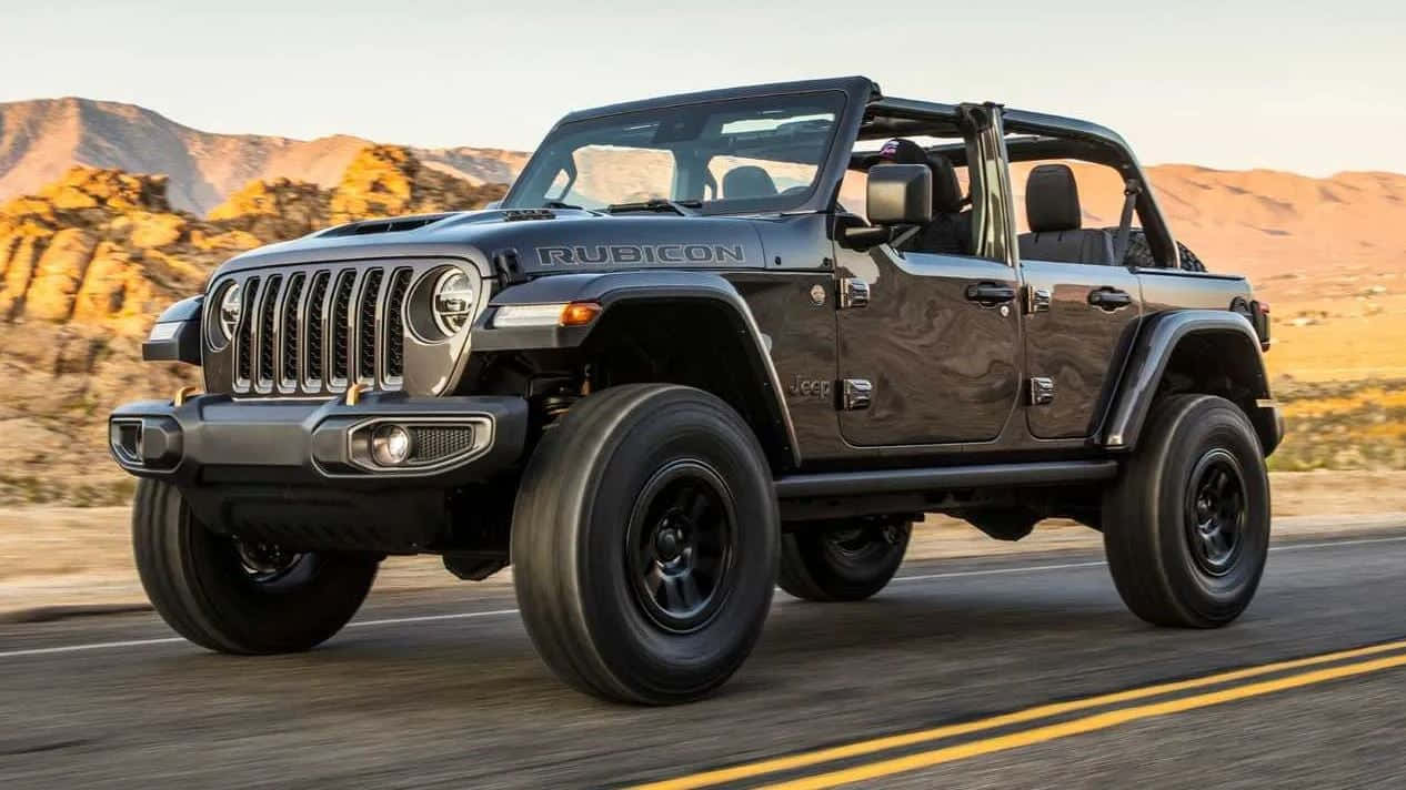 吉普 桌布 壁紙 — 一輛消光黑色的Jeep Wrangler Rubicon在沙漠公路上奔馳，岩石丘陵在溫暖的日落光芒中閃耀。