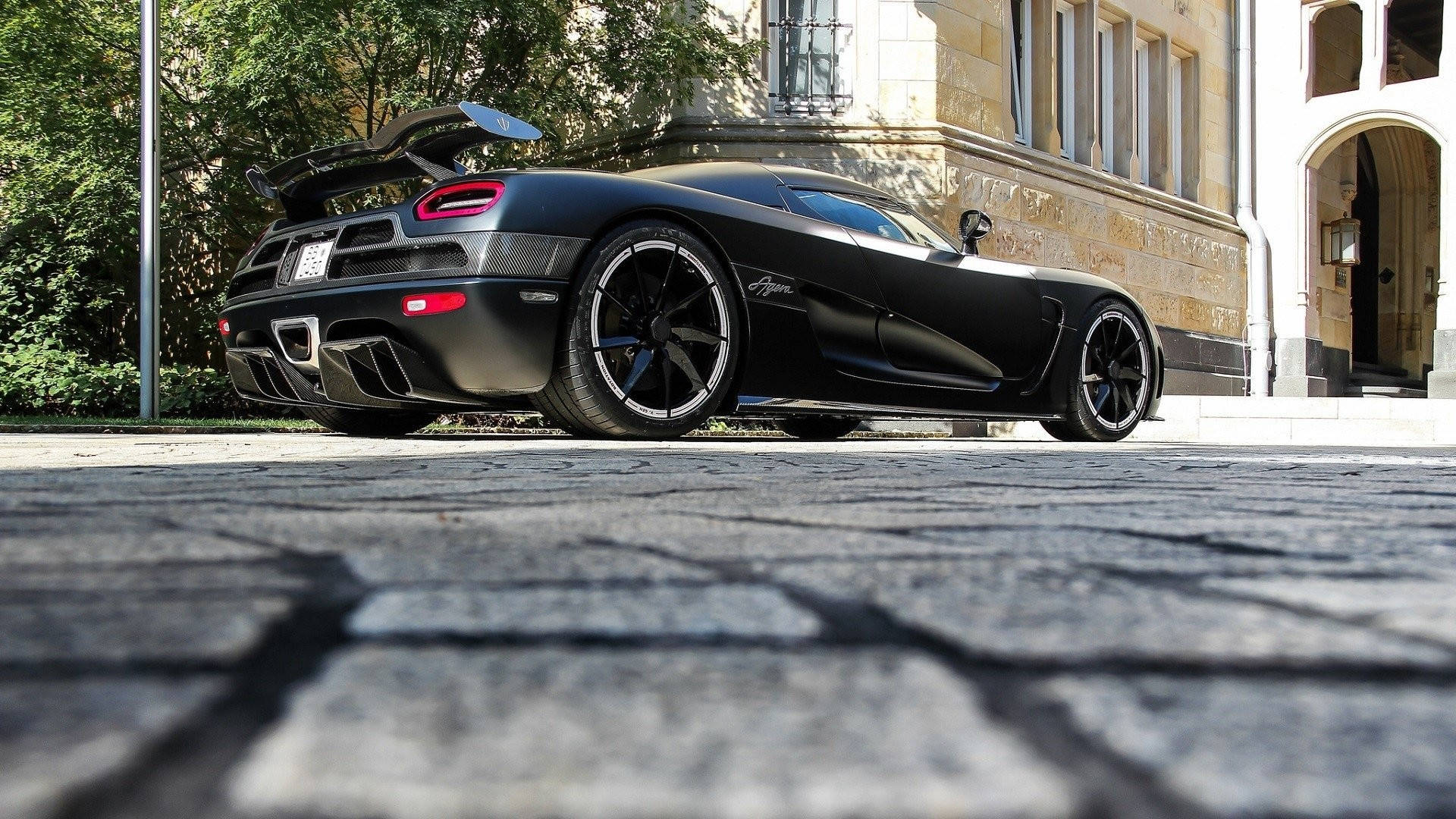 跑車 桌布 壁紙 — 一輛消光黑色的Koenigsegg Agera超跑停在鵝卵石路上，背景是宏偉的建築和樹木。