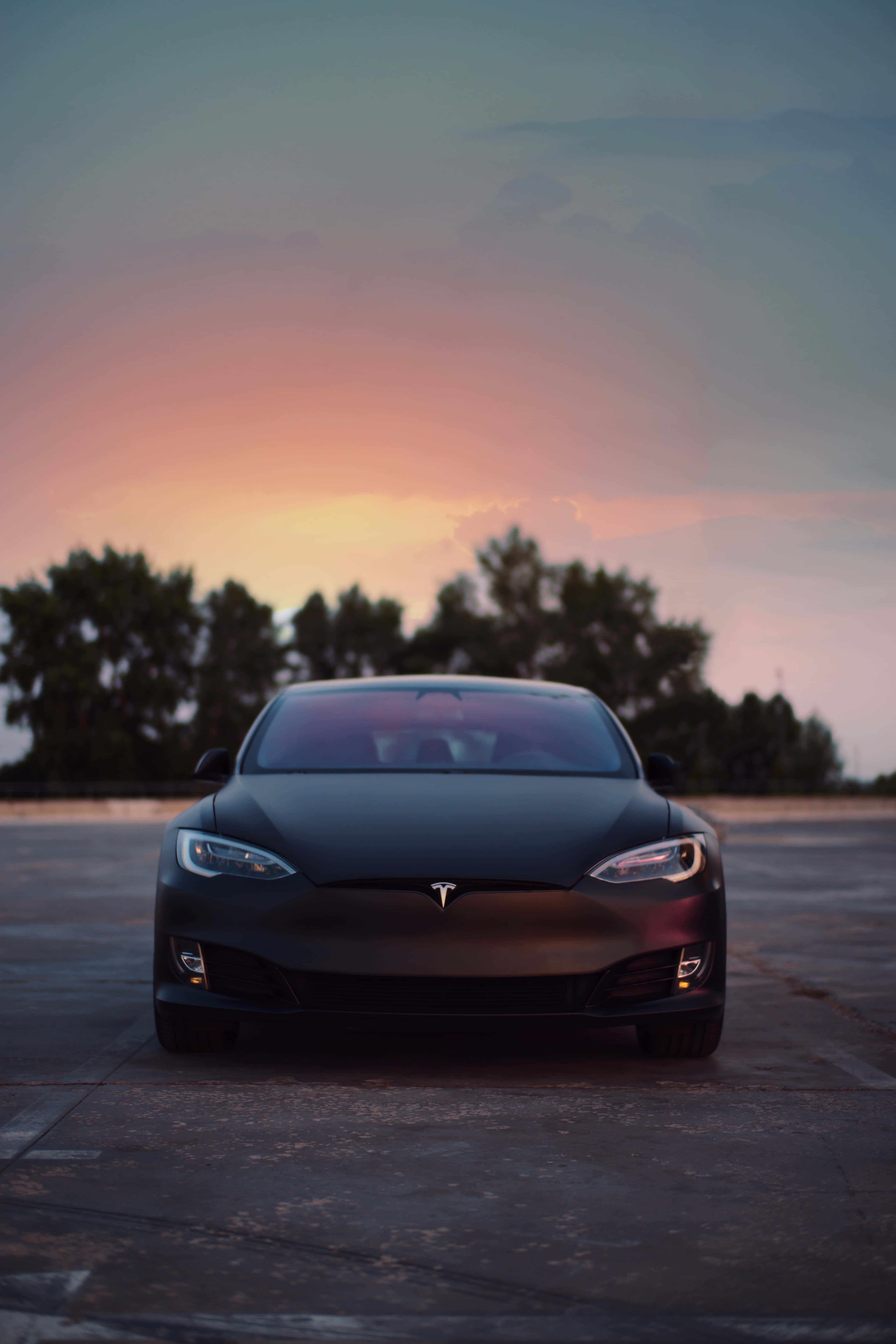特斯拉 桌布 壁紙 — 一輛消光黑色的Tesla Model S在鋪砌路面上向前行駛，柔和粉紅與橙色的日落色調在剪影樹木後方閃耀。