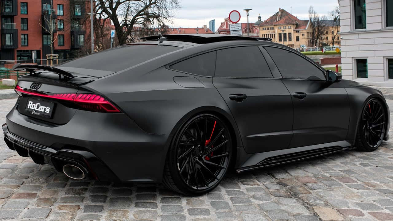 奧迪 桌布 壁紙 — 一輛消光黑色的奧迪RS7 Sportback，帶有紅色尾燈和制動卡鉗，停在城市環境中的鵝卵石街道上。