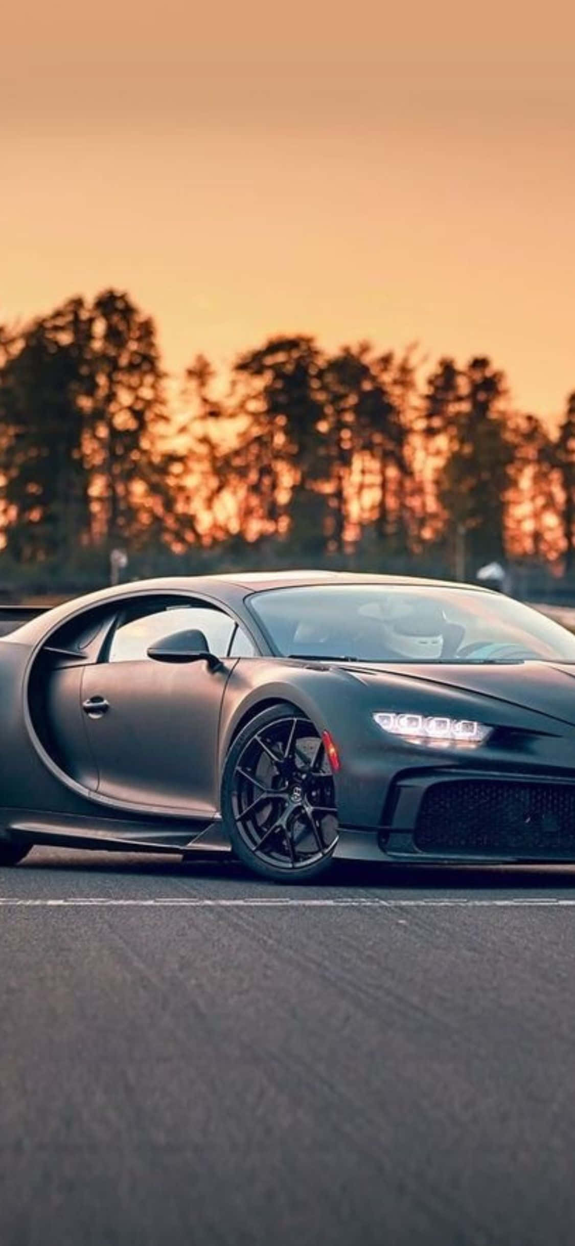 iPhone Xs Bugatti 桌布 壁紙 — 一輛消光黑色的布加迪Chiron停在柏油路上，背景是橘色的天空和剪影樹木的日落景色。
