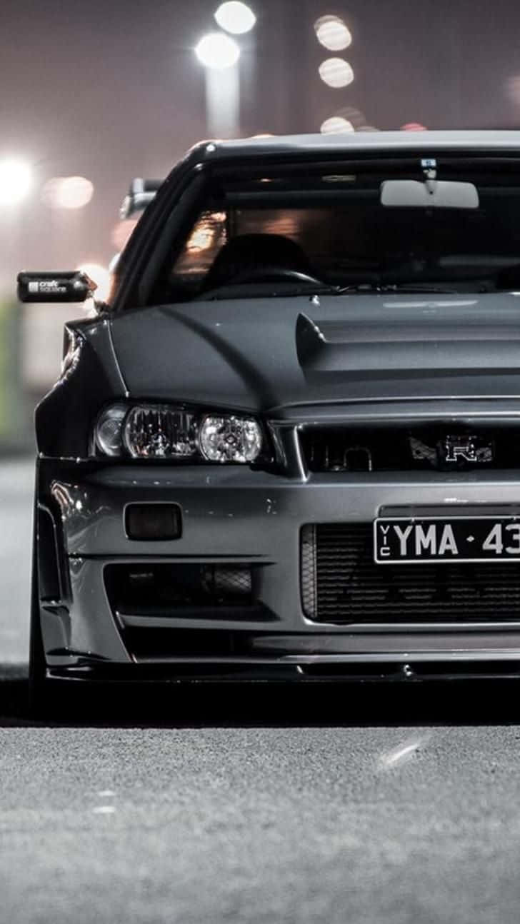 桌布 壁紙 — 一輛消光黑色的日產Skyline R34跑車在夜晚停泊在閃爍的路燈下，背景是模糊的城市景色。