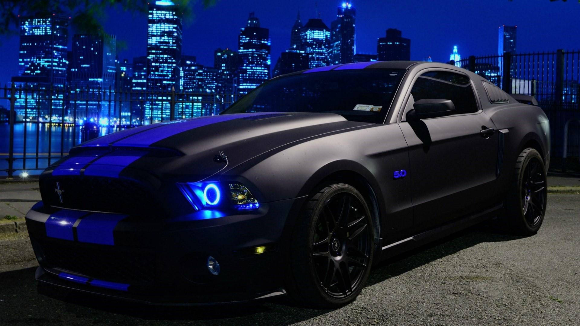 Ford Mustang 桌布 壁紙 — 一輛消光黑色的福特野馬，帶有發光的藍色點綴，在夜間停放在充滿活力的城市天際線前。