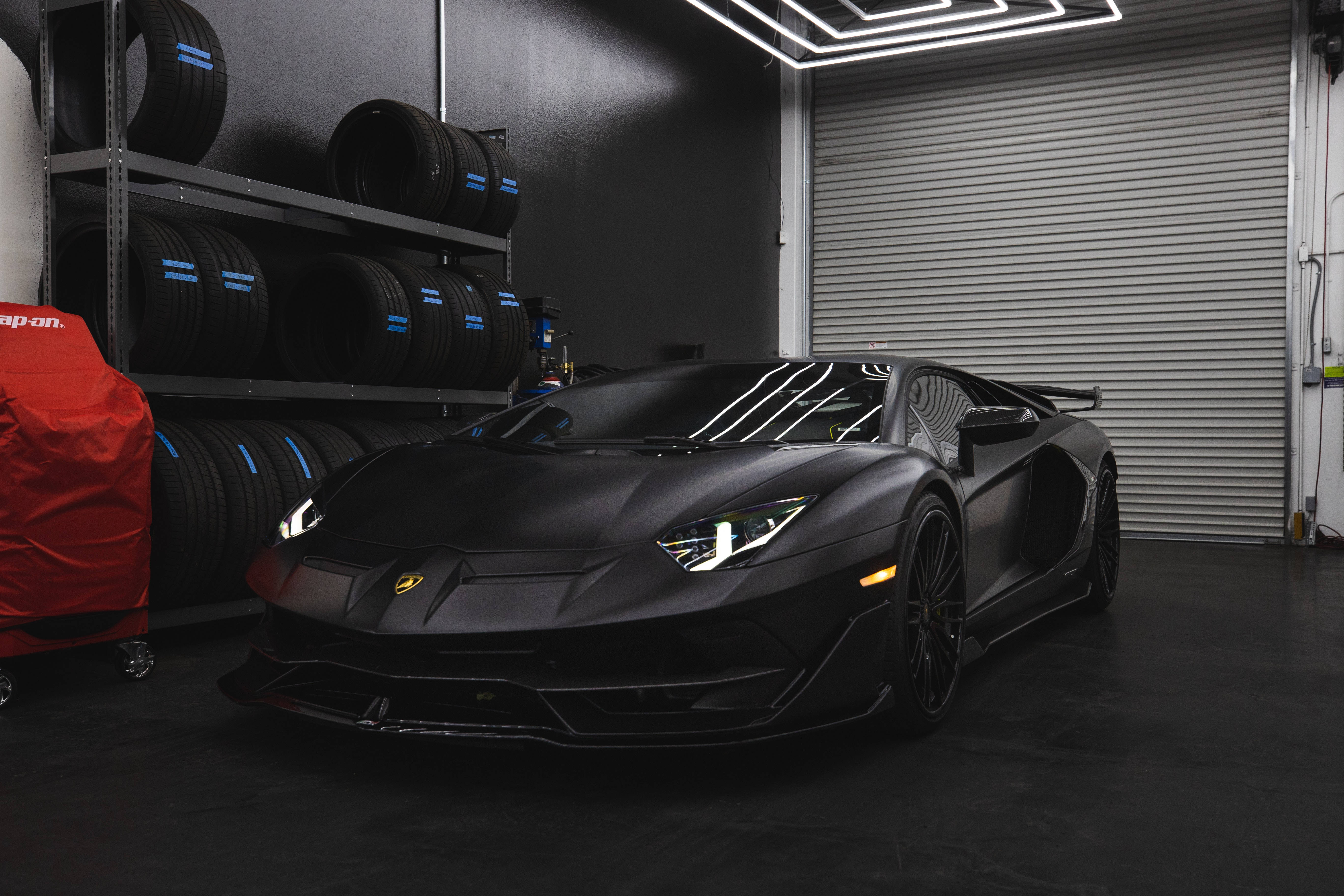 跑車 桌布 壁紙 — 一輛消光黑色的藍寶堅尼 Aventador SVJ 停放在一個時尚現代、配有堆疊輪胎和霓虹燈的車庫中。