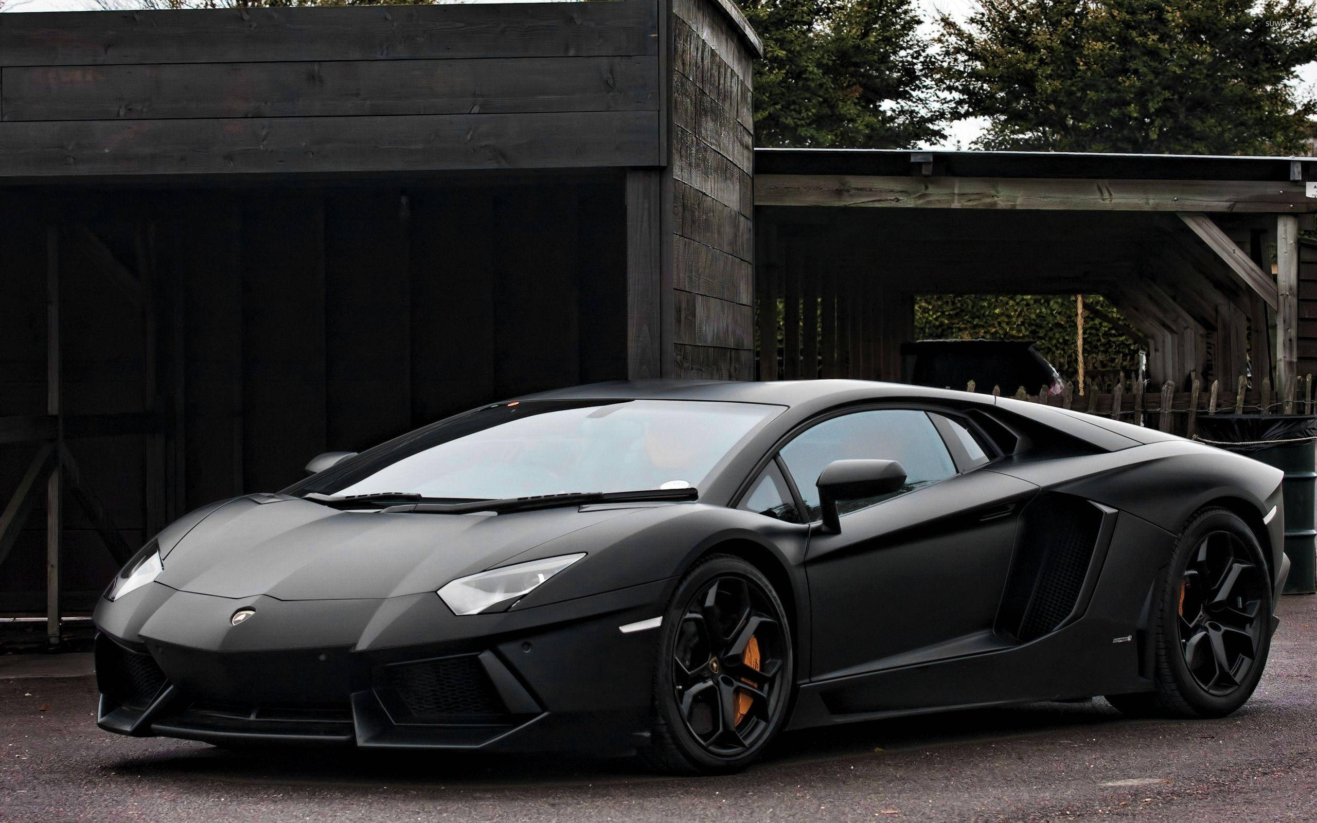 Lamborghini 4K 桌布 壁紙 — 一輛消光黑色的藍寶堅尼 Aventador 停放在質樸的木造結構前，橘色煞車卡鉗清晰可見。