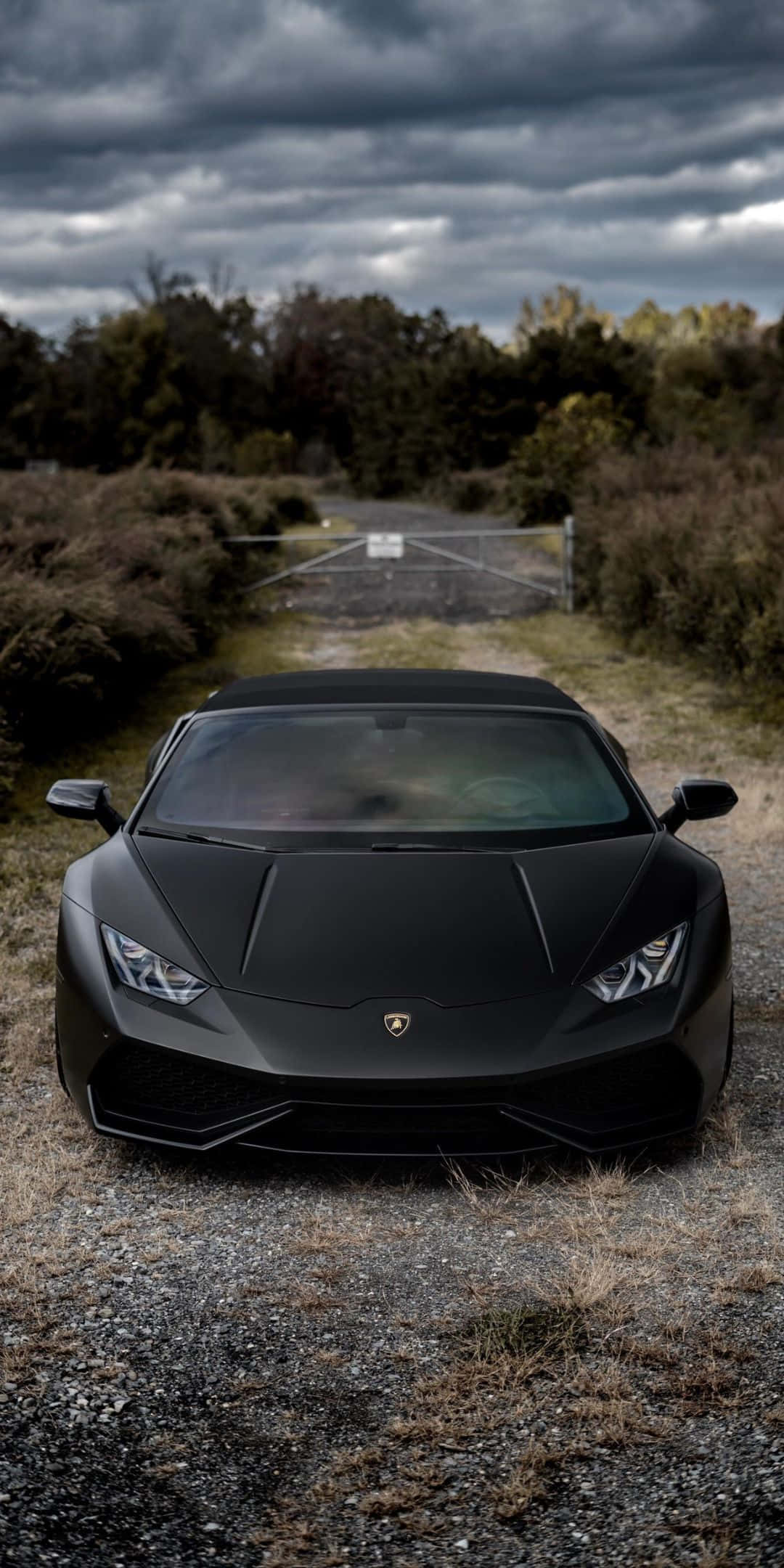 Pixel 3 Lamborghini 桌布 壁紙 — 一輛消光黑色的藍寶堅尼 Huracán 停在碎石路上，背景是暴風雨的天空和秋天的樹木。