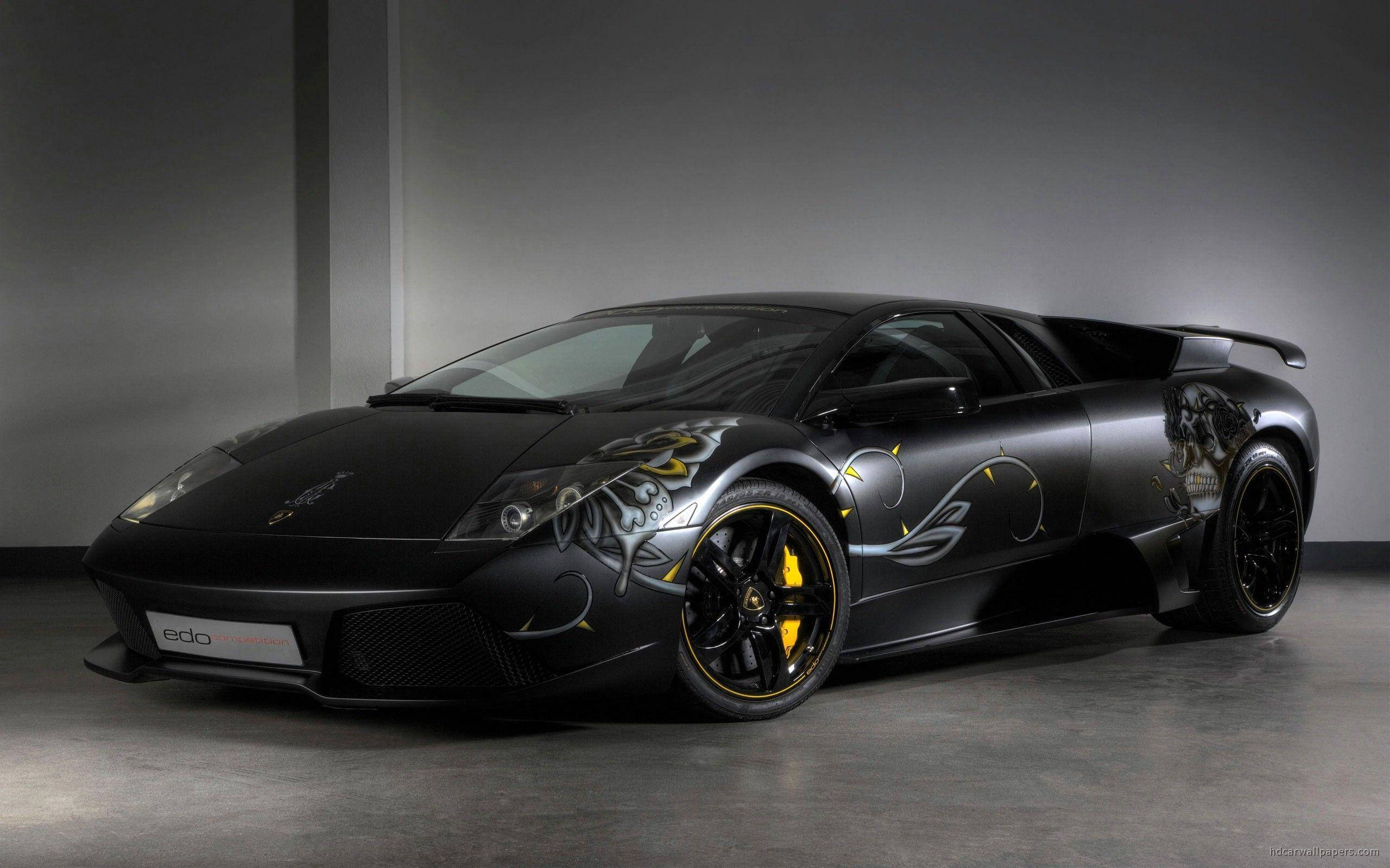 Lamborghini 4K 桌布 壁紙 — 一輛消光黑色的藍寶堅尼 Murciélago，帶有複雜的銀色和黃色部落圖案貼紙及骷髏圖案，停在簡約的工作室中。