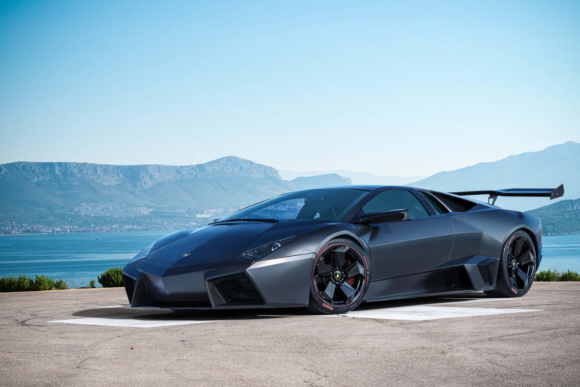 Lamborghini Reventón 桌布 壁紙 — 一輛消光黑色的藍寶堅尼Reventon跑車停泊在沿海公路上，背景是山脈和藍色海洋。