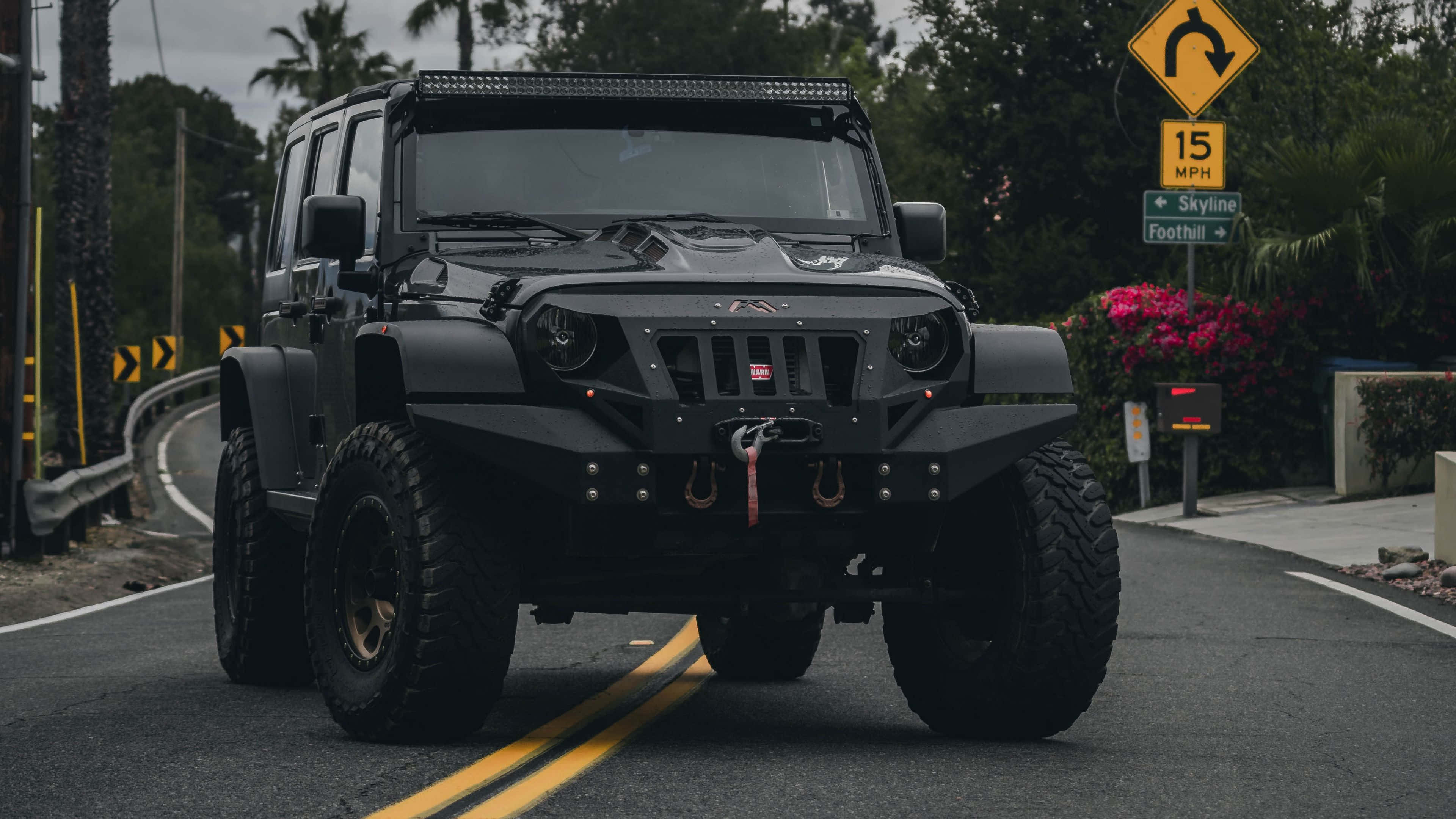 Jeep Cj 桌布 壁紙 — 一輛消光黑色的越野Jeep Wrangler配備超大輪胎，行駛在棕櫚樹和色彩繽紛的花朵環繞的蜿蜒道路上。