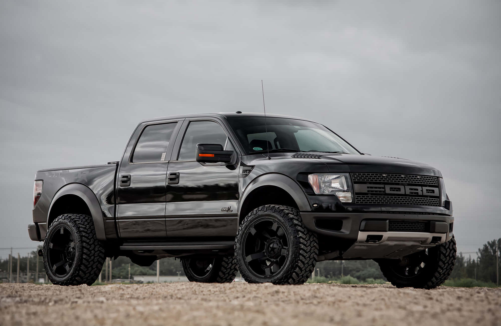 福特 桌布 壁紙 — 一輛消光黑色福特F-150 Raptor在陰天的天空下，配備堅固輪胎和升高懸吊，高高立在碎石上。