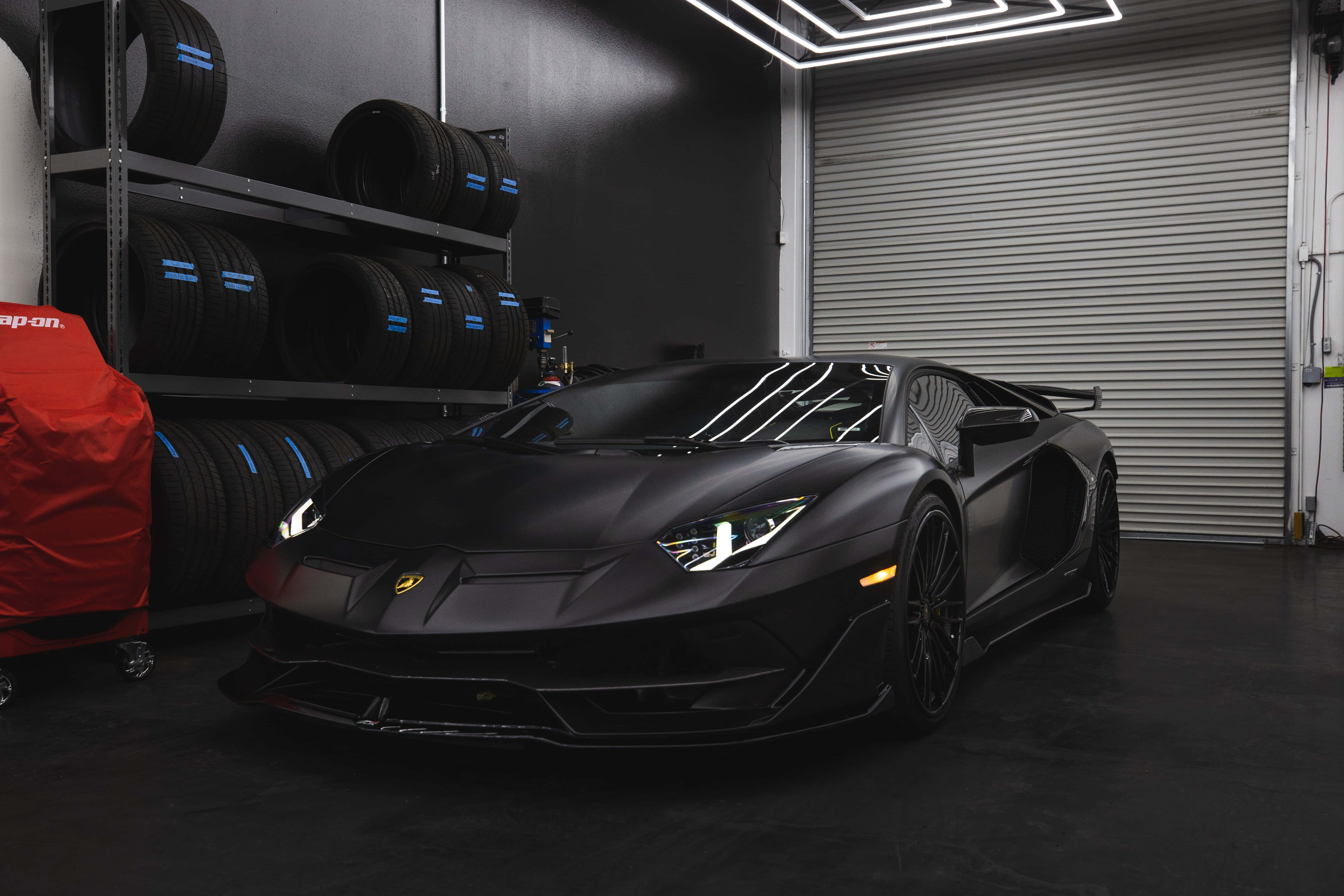 霧面黑色 桌布 壁紙 — 一輛消光黑色藍寶堅尼 Aventador SVJ 停放在一個時尚現代、帶有堆疊輪胎和霓虹燈的車庫中。