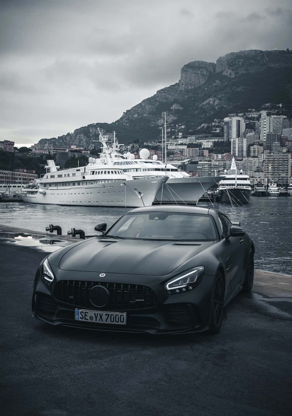 Mercedes 桌布 壁紙 — 一輛消光黑色跑車停泊在港口碼頭，背景有豪華遊艇和懸崖城市，天色陰沉。