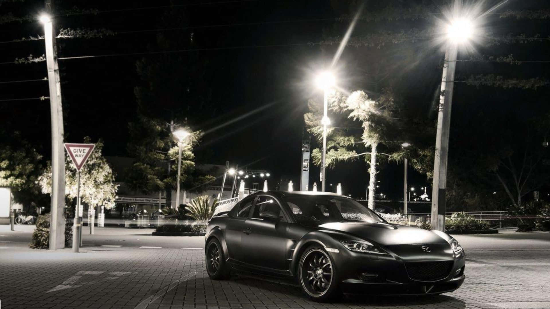 Mazda Rx-8 桌布 壁紙 — 一輛消光黑色跑車在寧靜的夜間城市廣場上，在明亮的街燈下閃閃發光，周圍環繞著樹木和標誌。