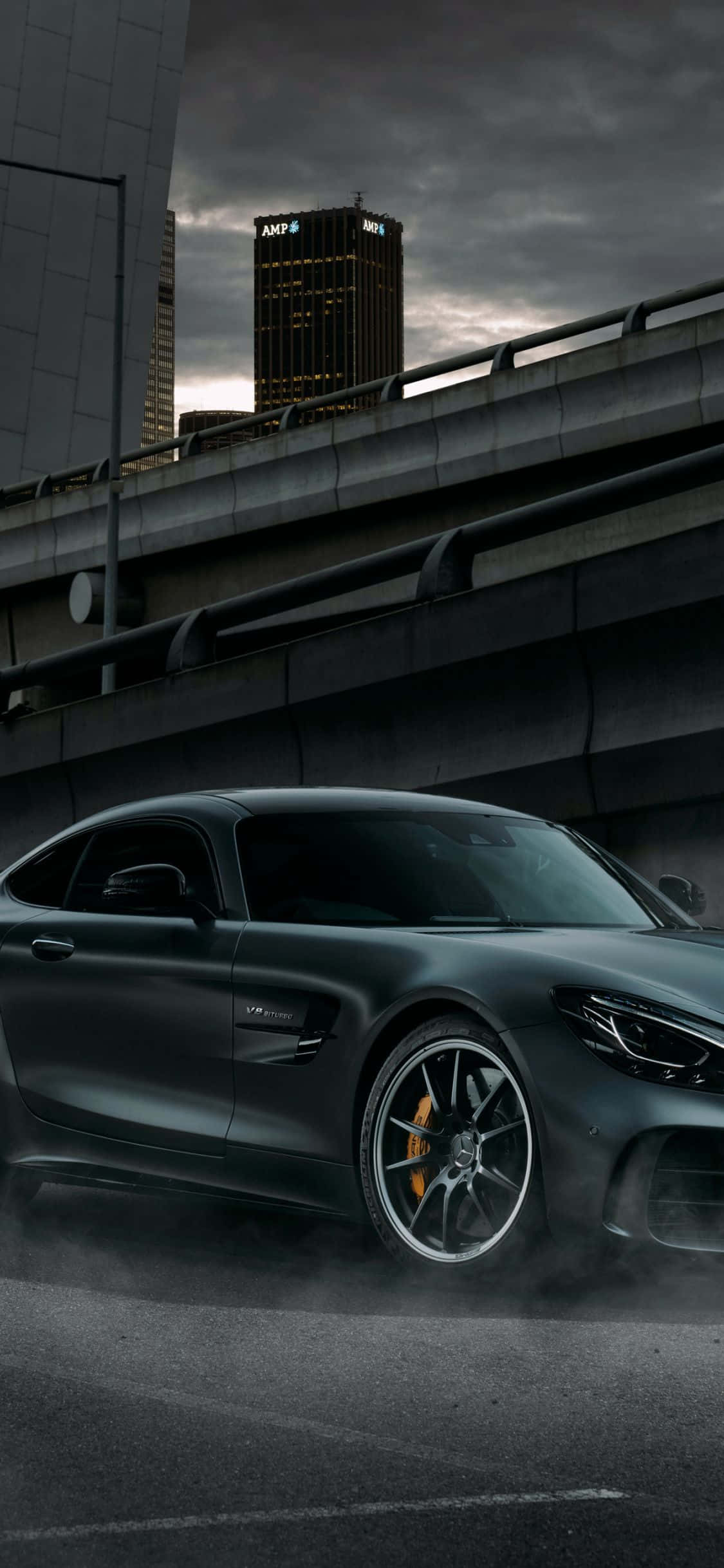 Mercedes 桌布 壁紙 — 一輛消光黑色跑車在潮濕的城市高架橋上揚起灰塵，背景是暴風雨的天空和燈火通明的摩天大樓。