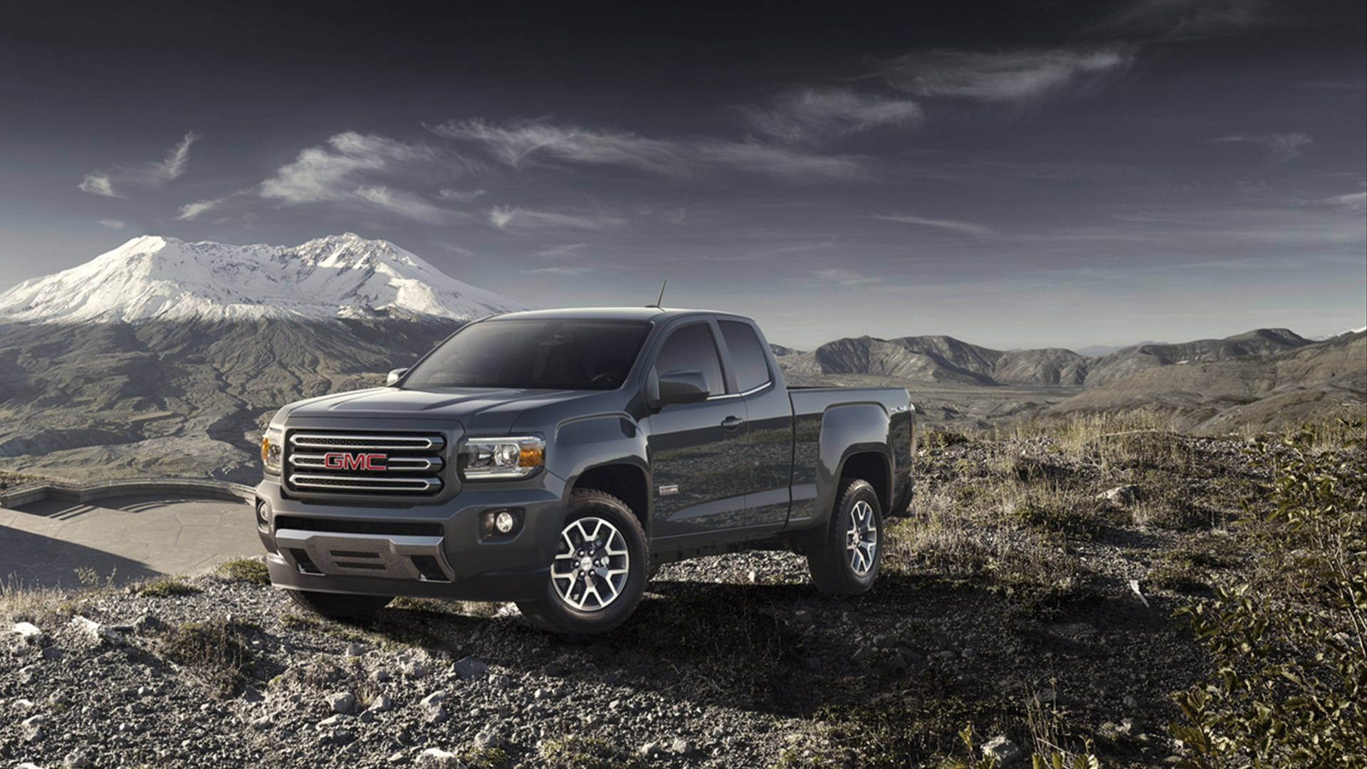Gmc 桌布 壁紙 — 一輛深灰色GMC Canyon皮卡車停放在岩石地形上，背景是雲層密布的天空下的一座雪山。
