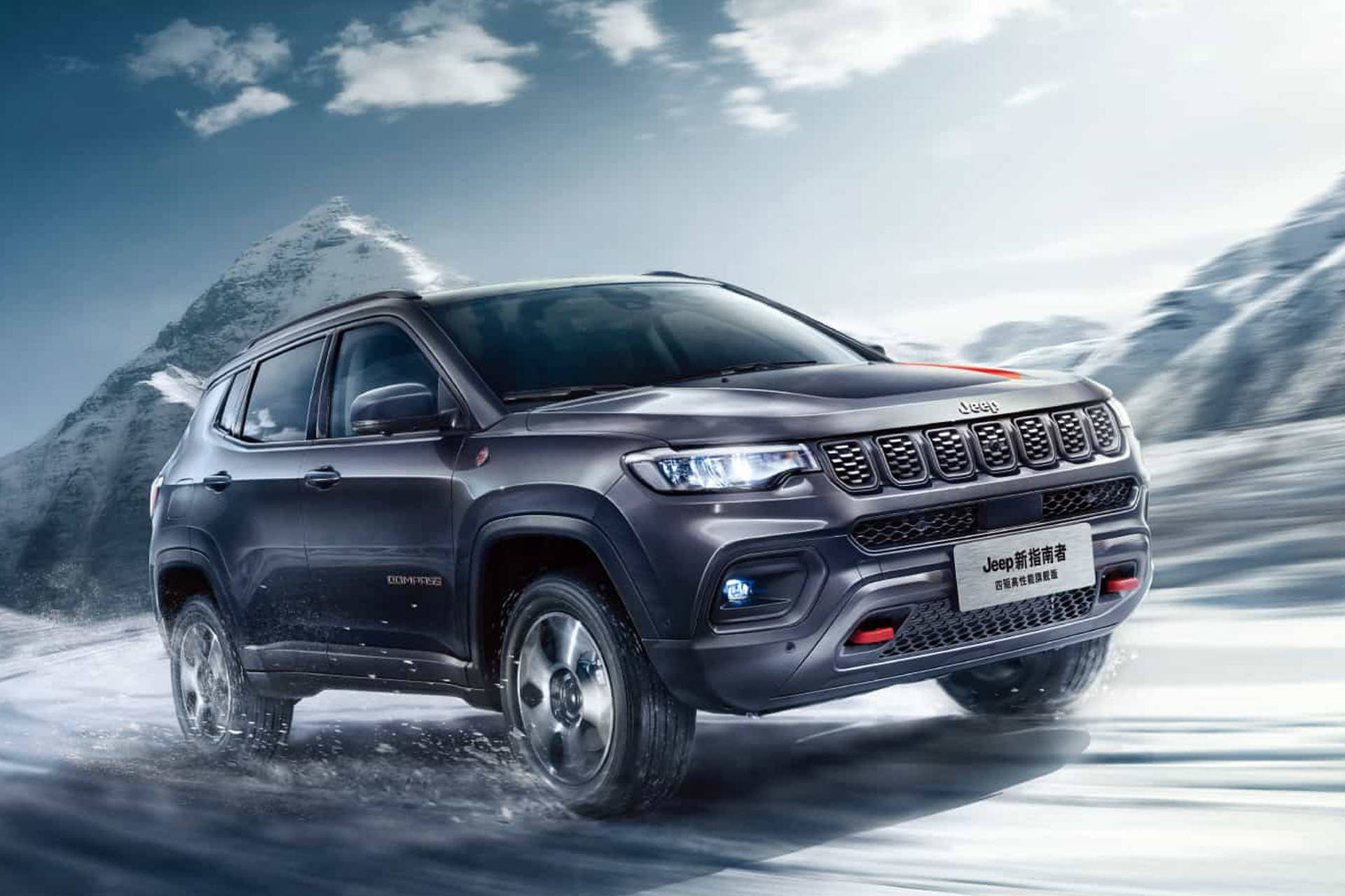  壁紙 — 一輛深灰色Jeep Compass SUV在雪地中行駛，車輪濺起雪花，背景是雲天下積雪覆蓋的山脈。