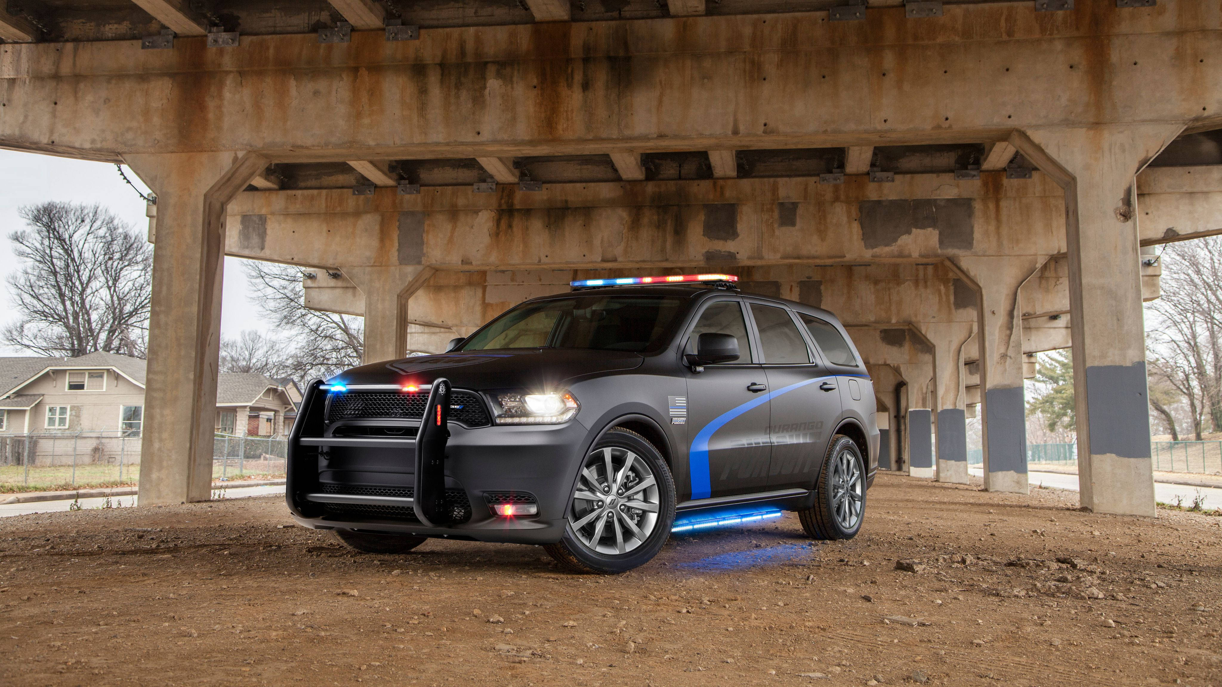 Dodge 桌布 壁紙 — 一輛深灰色警用SUV，閃爍著藍色和紅色的燈光，停泊在風化混凝土高架橋下的裸露地面上。