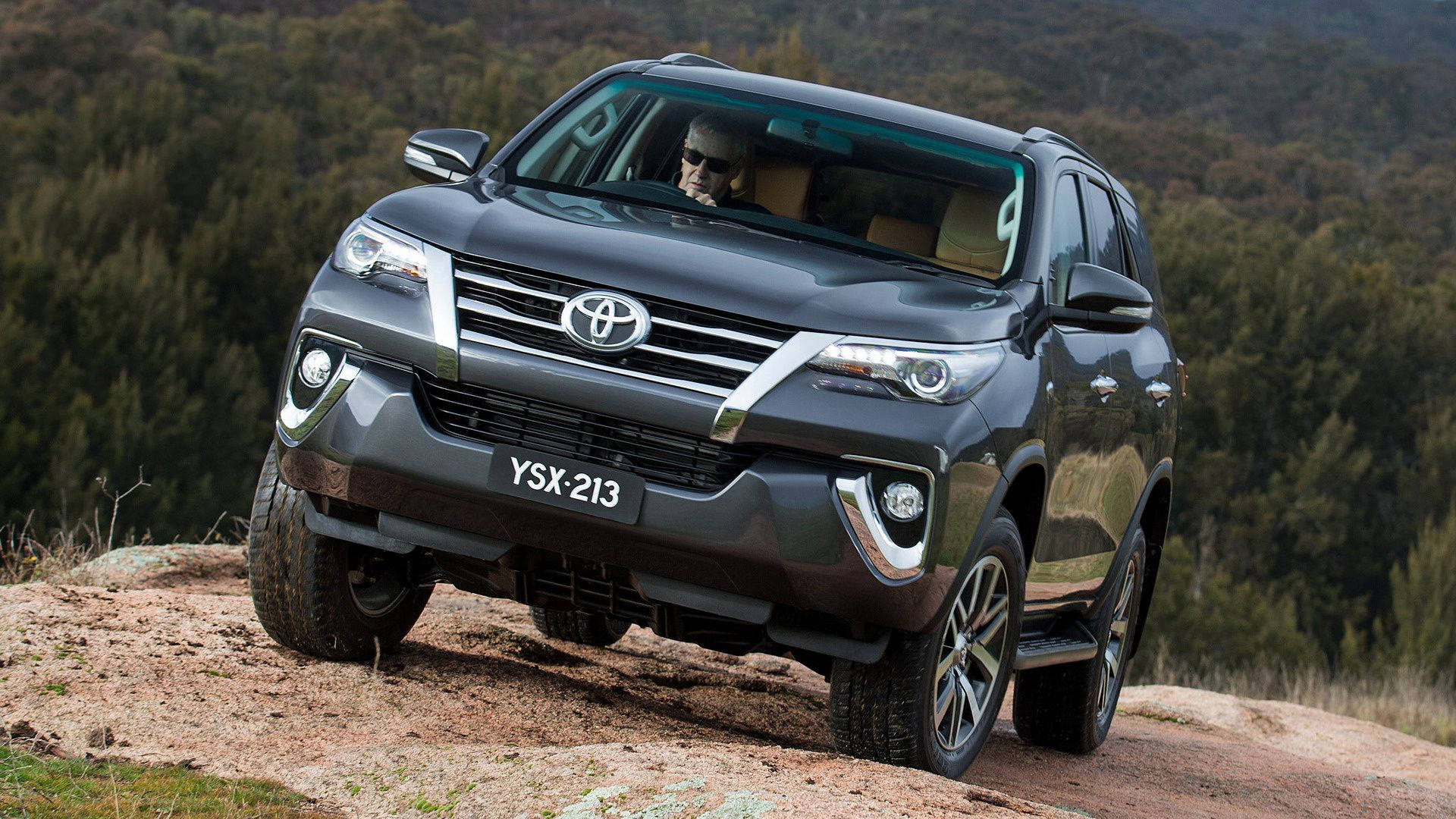 Outdoor 桌布 壁紙 — 一輛深灰色豐田 Fortuner SUV 自信地行駛在崎嶇的碎石地形上，背景是樹木繁茂的山坡。