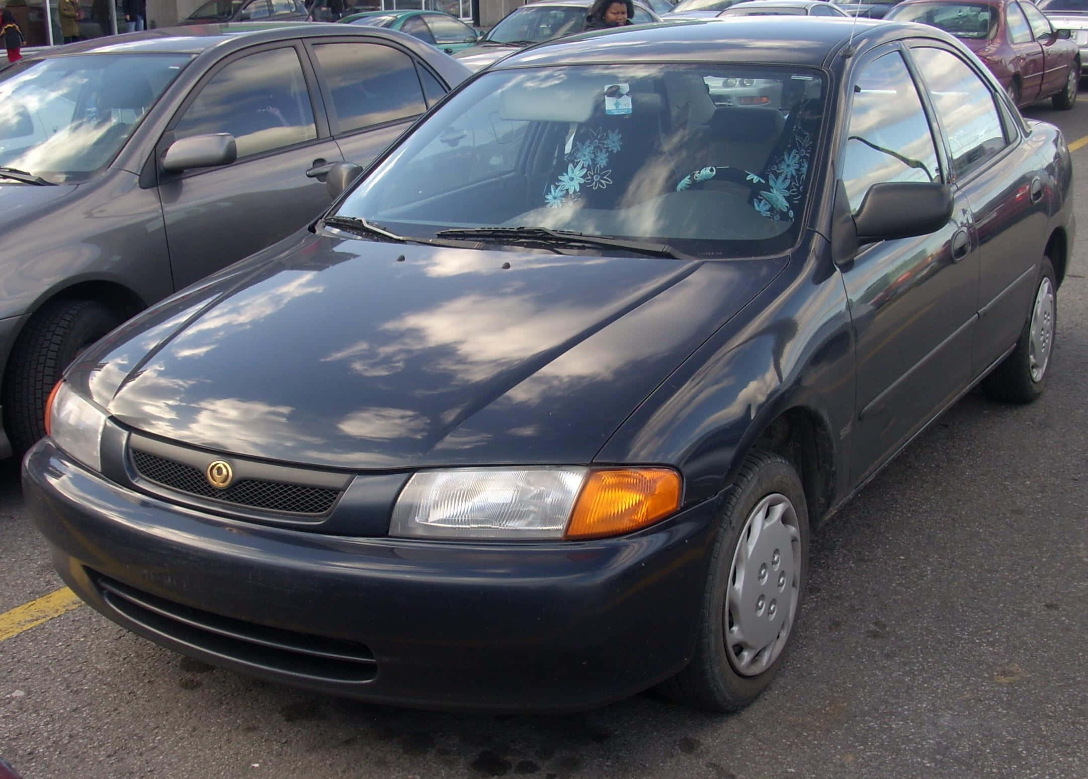 Mazda Protege 桌布 壁紙 — 一輛深灰色轎車停放在停車場，引擎蓋上反射著天空，擋風玻璃上有花卉貼紙。
