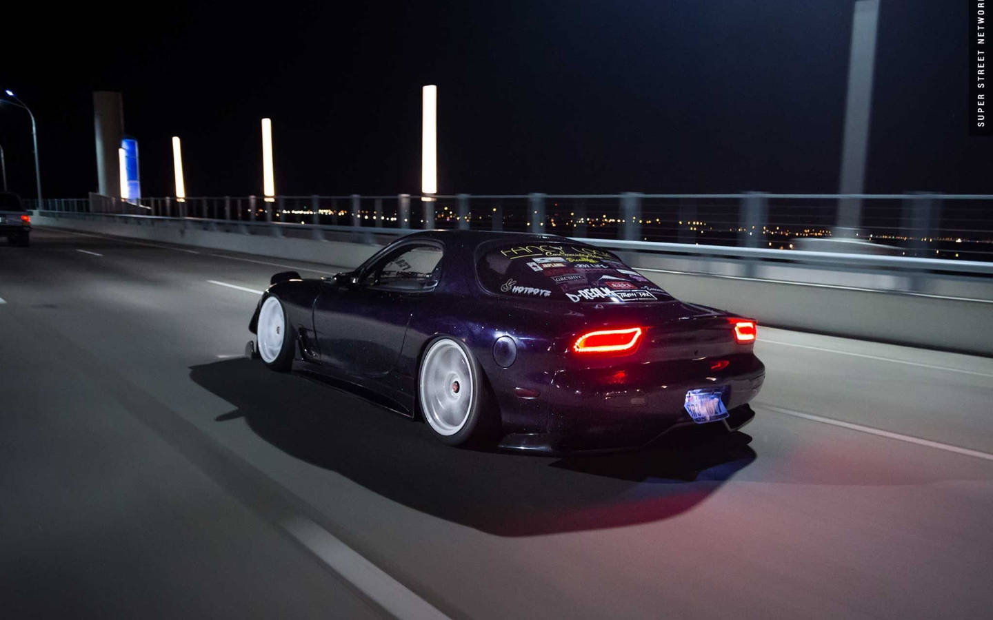 Mazda 桌布 壁紙 — 一輛深紫色改裝跑車在夜間行駛於燈火通明的橋上，遠處城市燈光閃爍。