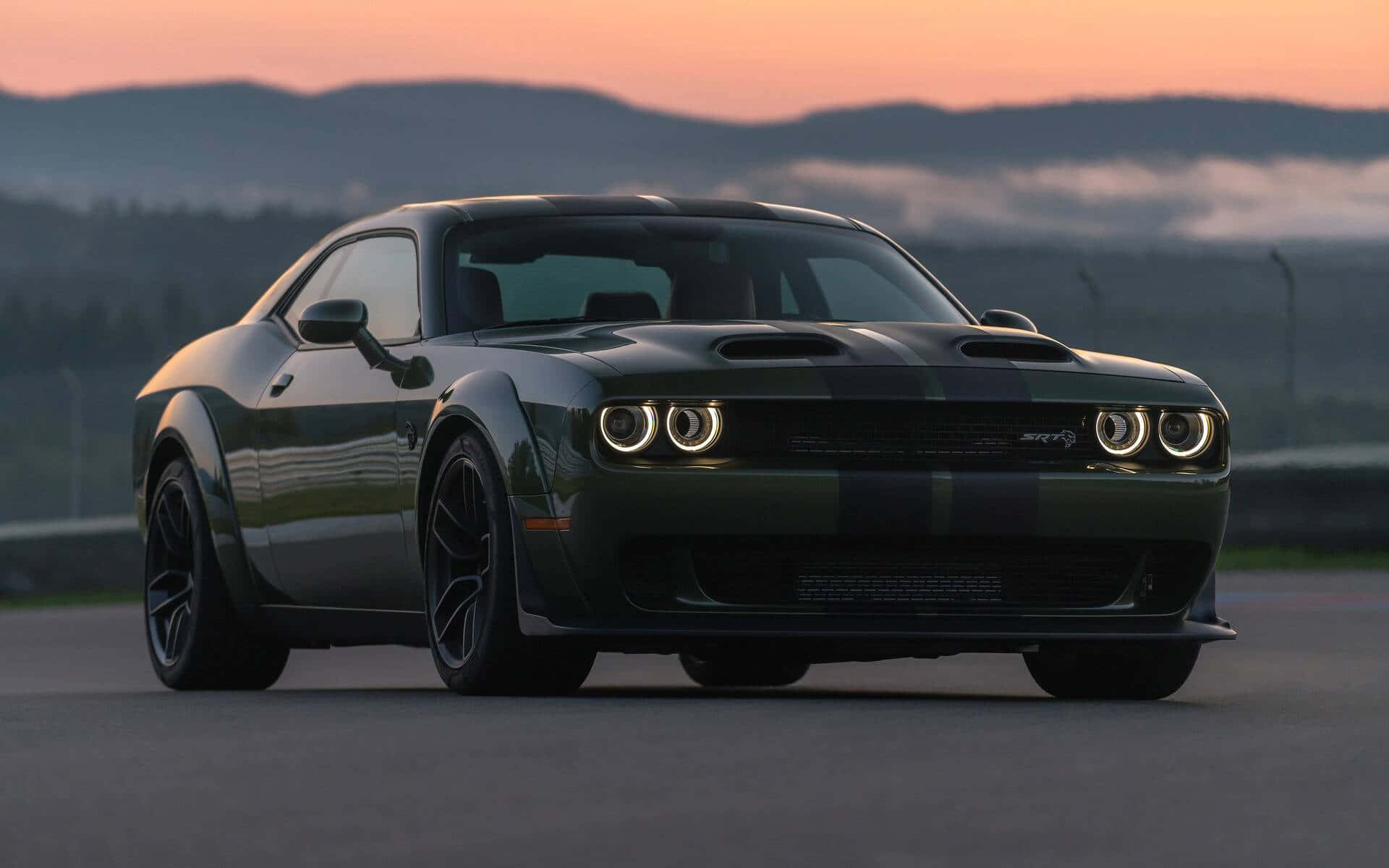 道奇 桌布 壁紙 — 一輛深綠色Dodge Challenger SRT Hellcat帶有黑色賽車條紋，停在日落時分的賽道上，背景是霧氣繚繞的山脈。