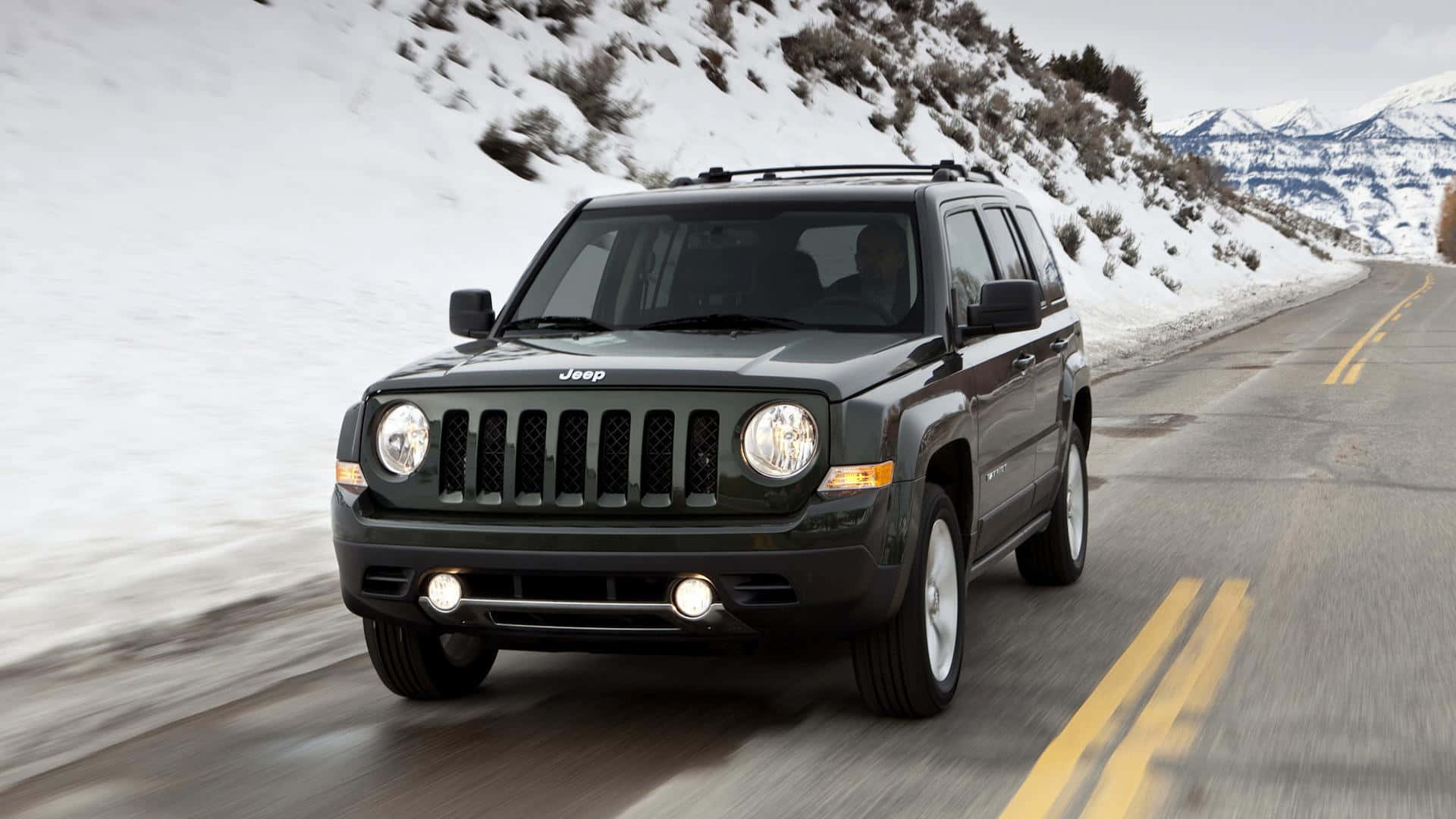 Jeep Patriot 桌布 壁紙 — 一輛深綠色Jeep Patriot在蜿蜒的山路上行駛，穿越積雪的地形，遠方有山峰。
