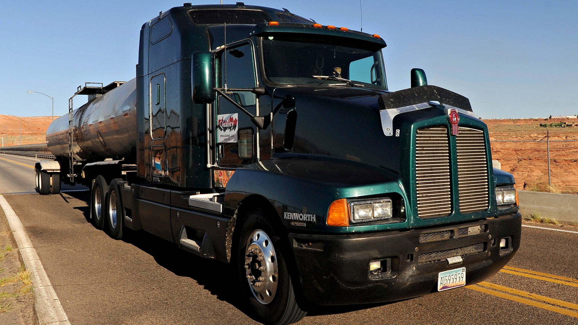 Kenworth 桌布 壁紙 — 一輛深綠色Kenworth油罐車在晴朗藍天下行駛於沙漠公路，背景有紅色岩層。