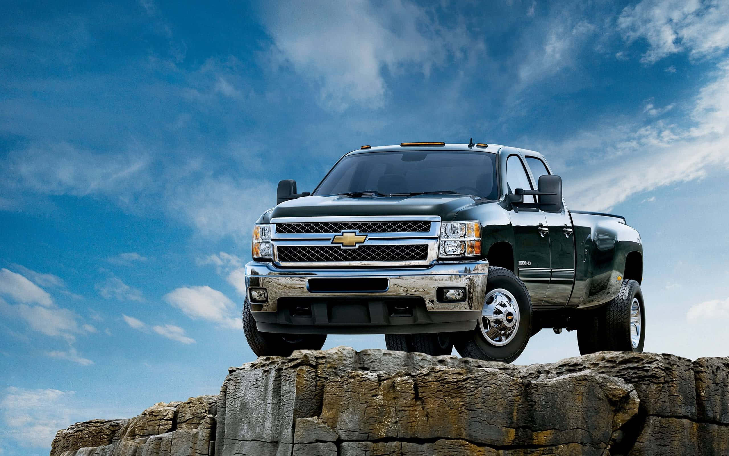 chevy 桌布 壁紙 — 一輛深綠色的 Chevrolet Silverado 皮卡車強而有力地停在崎嶇岩石懸崖上，背景是晴朗藍天和零星白雲。