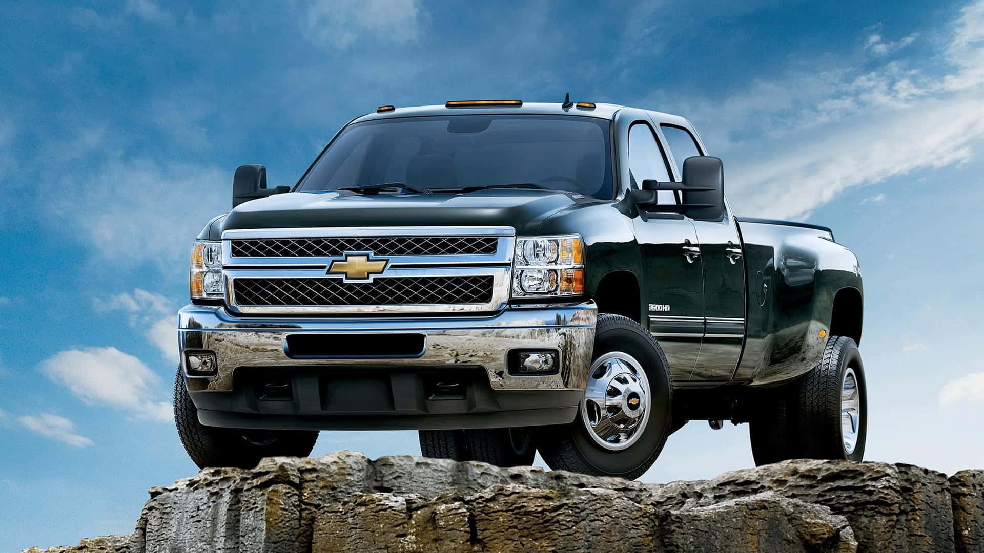 攝影 桌布 壁紙 — 一輛深綠色的 Chevrolet Silverado  pickup truck 自豪地矗立在崎嶇岩石上，天空湛藍，白雲點綴。