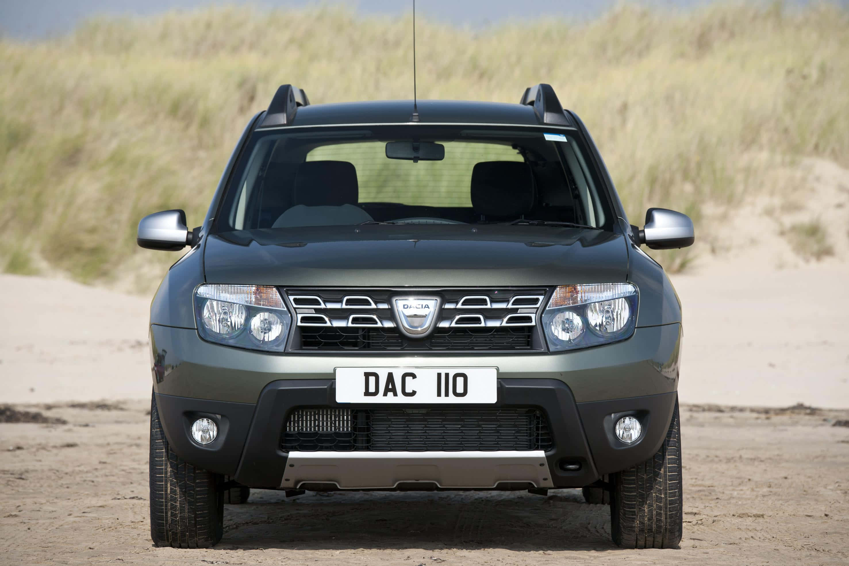 Dacia 桌布 壁紙 — 一輛深綠色的Dacia Duster SUV在明亮的日光下，面向沙灘，背後有沙丘和乾草。