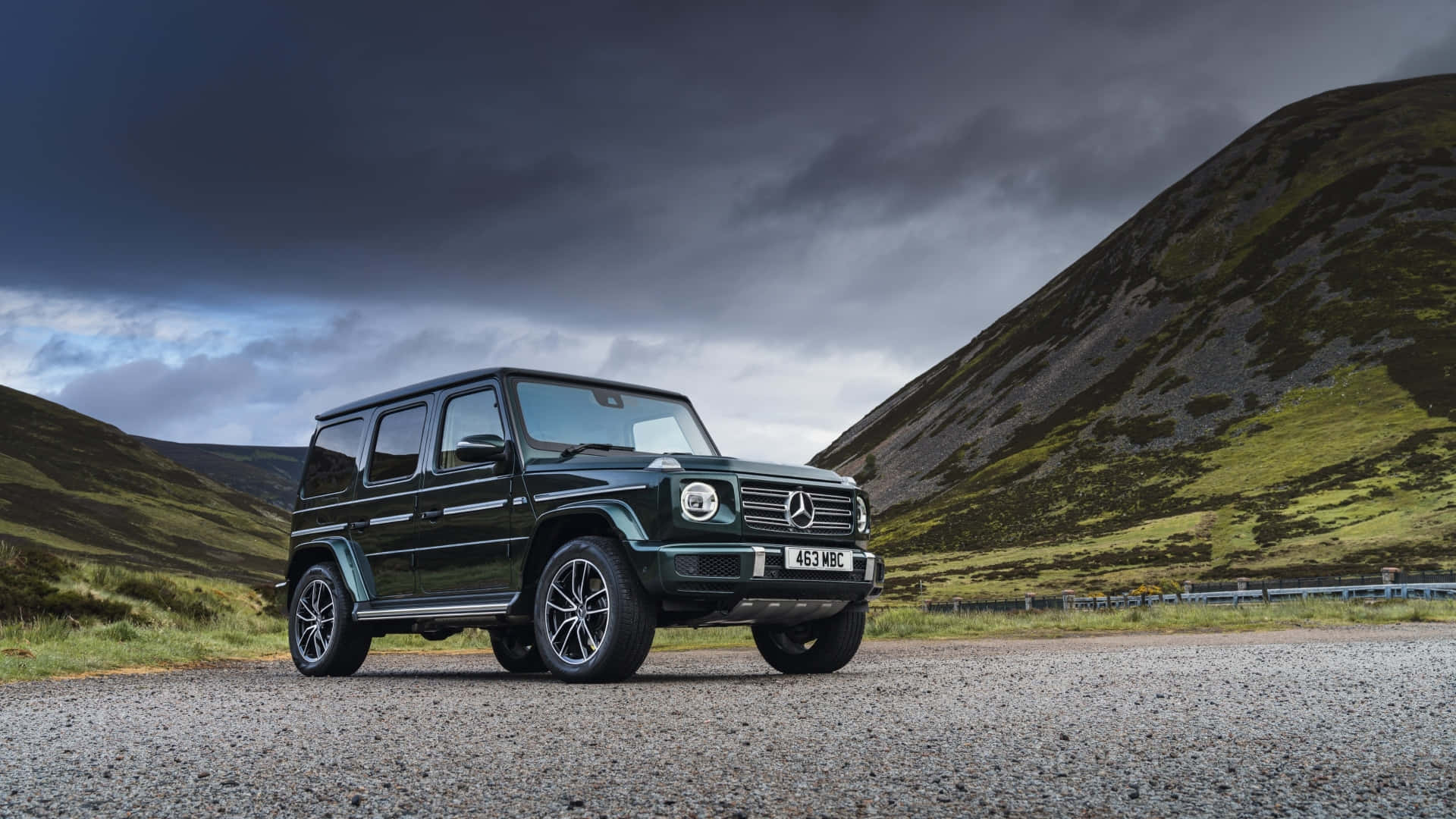 Mercedes Car 4K 桌布 壁紙 — 一輛深綠色賓士 G-Class SUV 停放在碎石路上，背景是起伏的綠色山丘，上方籠罩著威脅性的暴風雲。