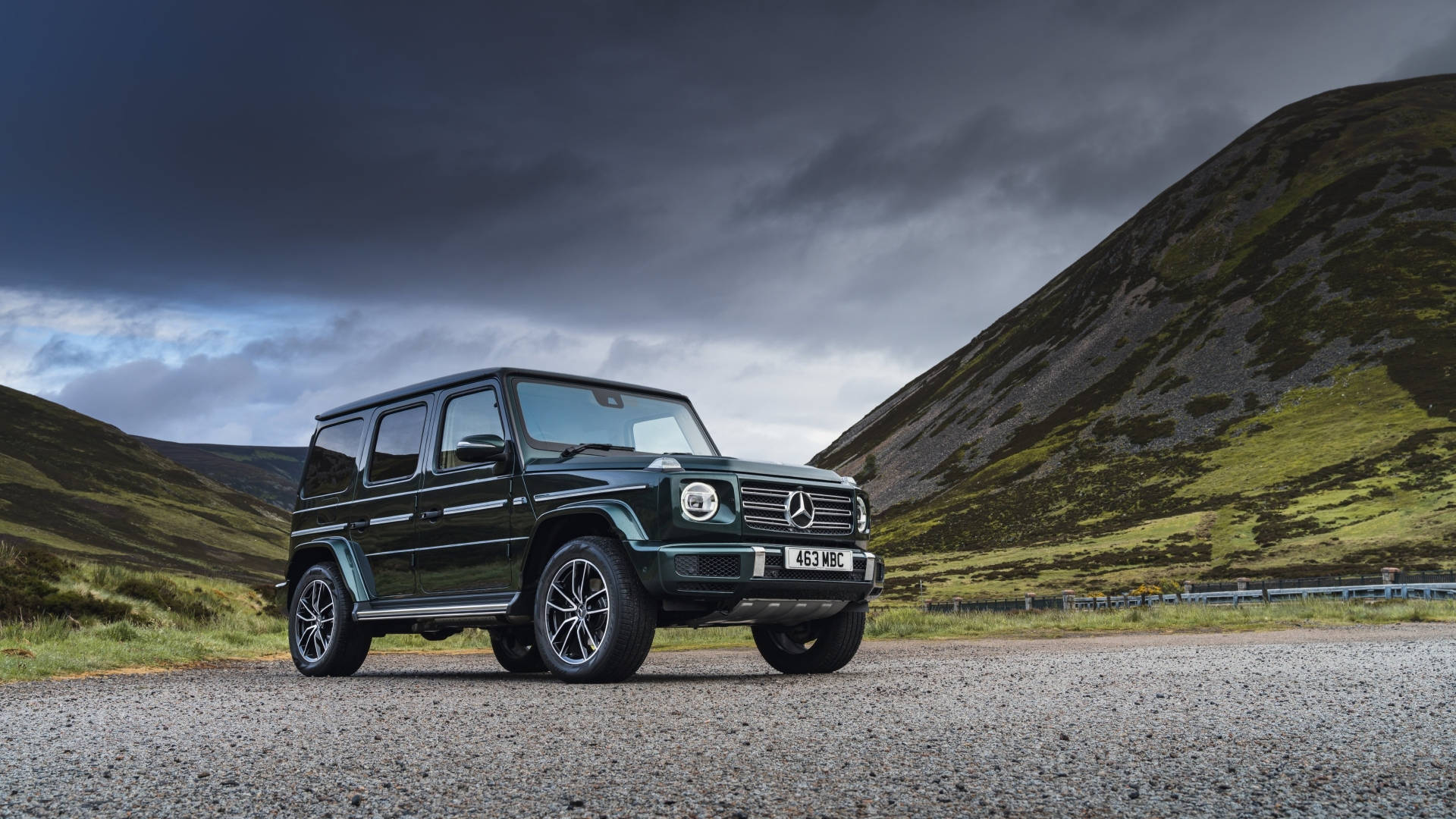 山景 桌布 壁紙 — 一輛深綠色賓士 G-Class SUV 在礫石路上，在翻滾的綠色山丘背景下，傲然矗立於威脅性的暴風雲之下。