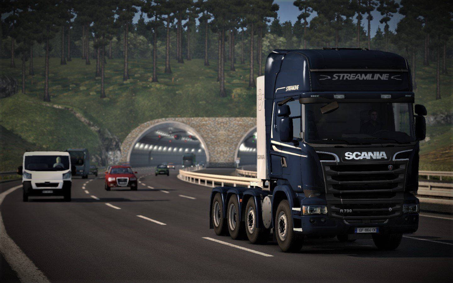 American Truck Simulator 桌布 壁紙 — 一輛深藍色 Scania Streamline R730 卡車帶領車隊在蜿蜒的山區高速公路上行駛，接近一個兩旁是茂密松林的石隧道。