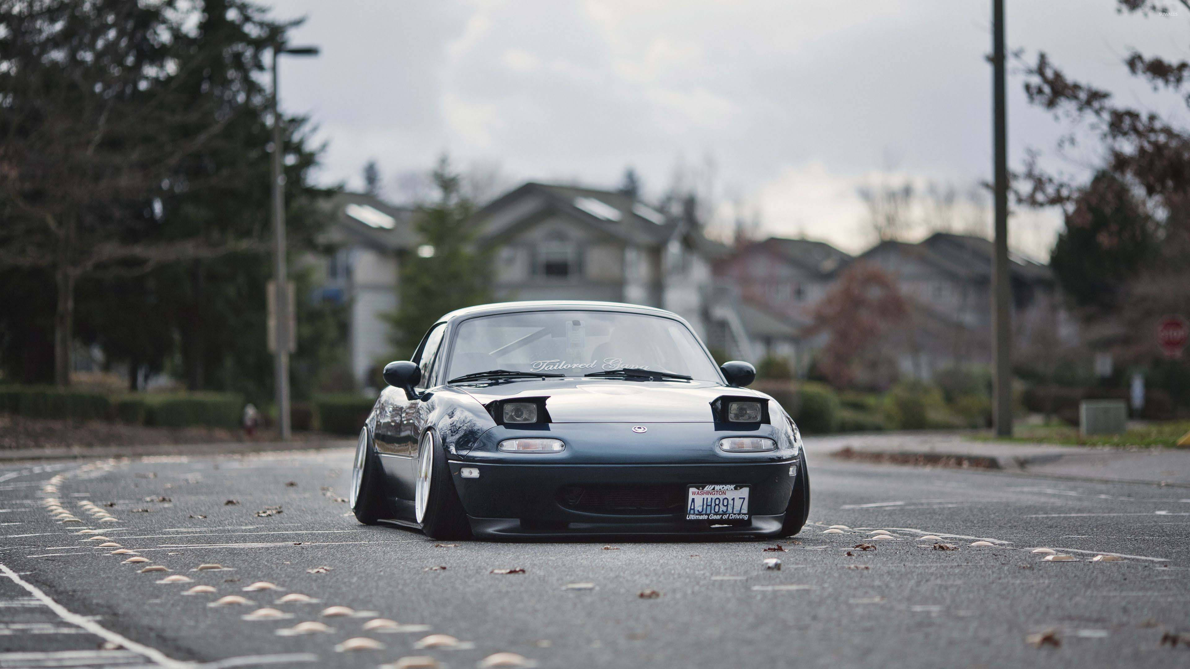 JDM 桌布 壁紙 — 一輛深藍色Mazda Miata，降低懸吊，停在散落秋葉的郊區街道中央。